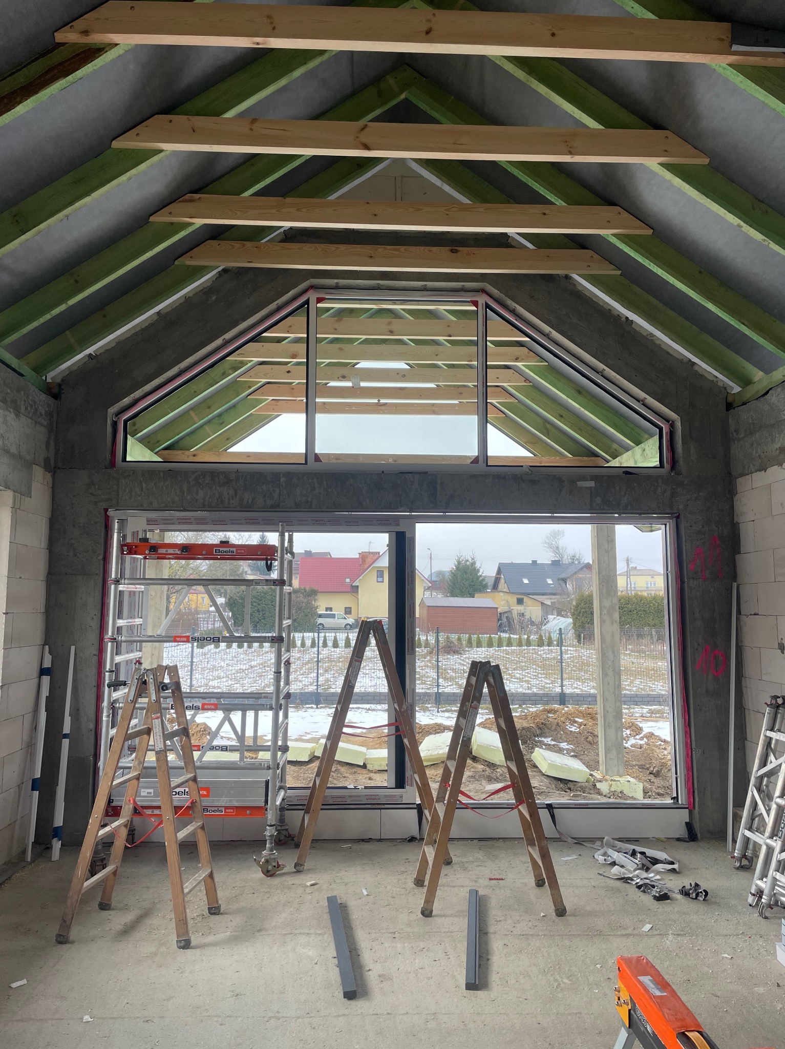 Wnętrze surowego budynku z zamontowanym oknem w kształcie trapezu, widoczne rusztowania i elementy konstrukcji dachu z drewna. Na zewnątrz krajobraz zimowy.