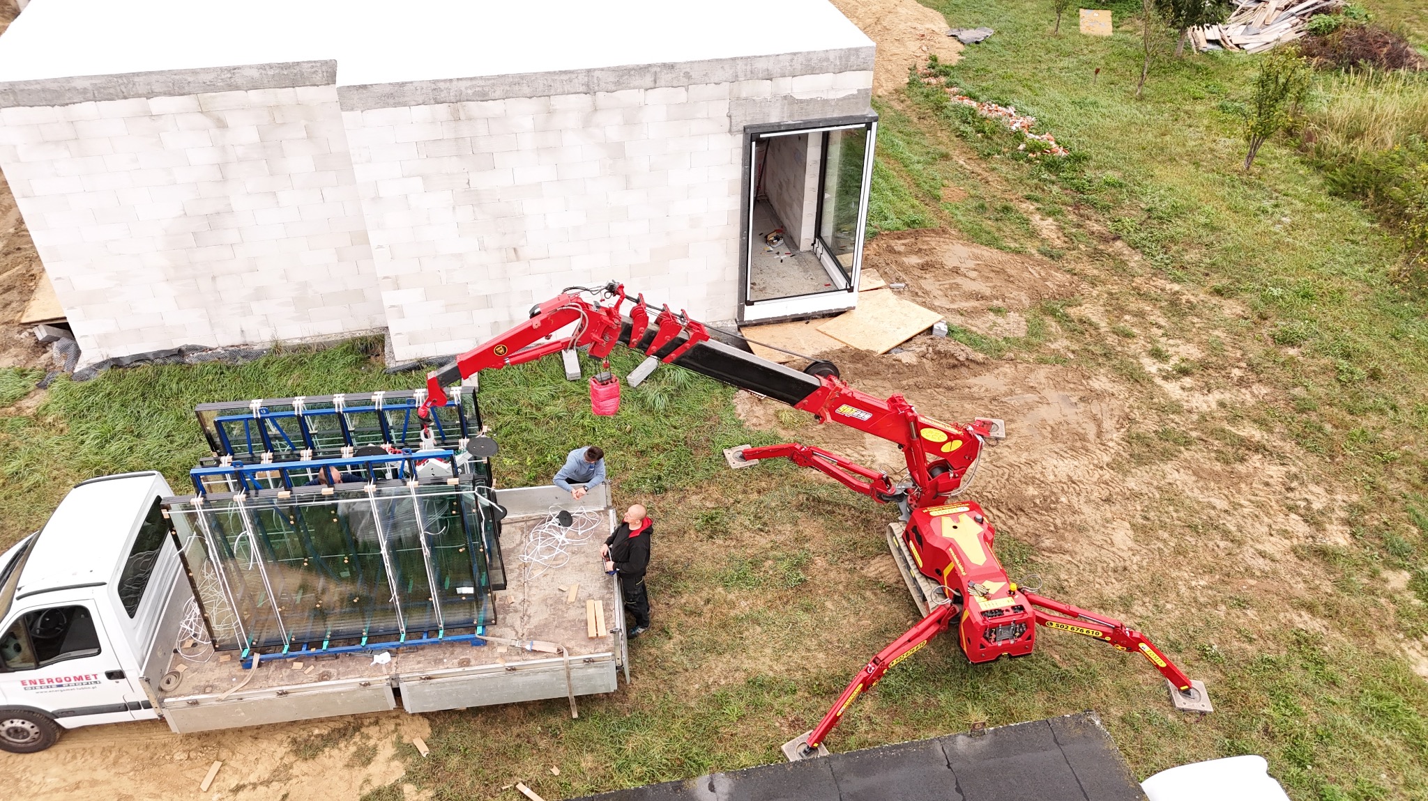 Montaż okien z użyciem dźwigu na budowie. Okna na samochodzie dostawczym, pracownicy nadzorują proces. Widok z góry na budynek w trakcie budowy.