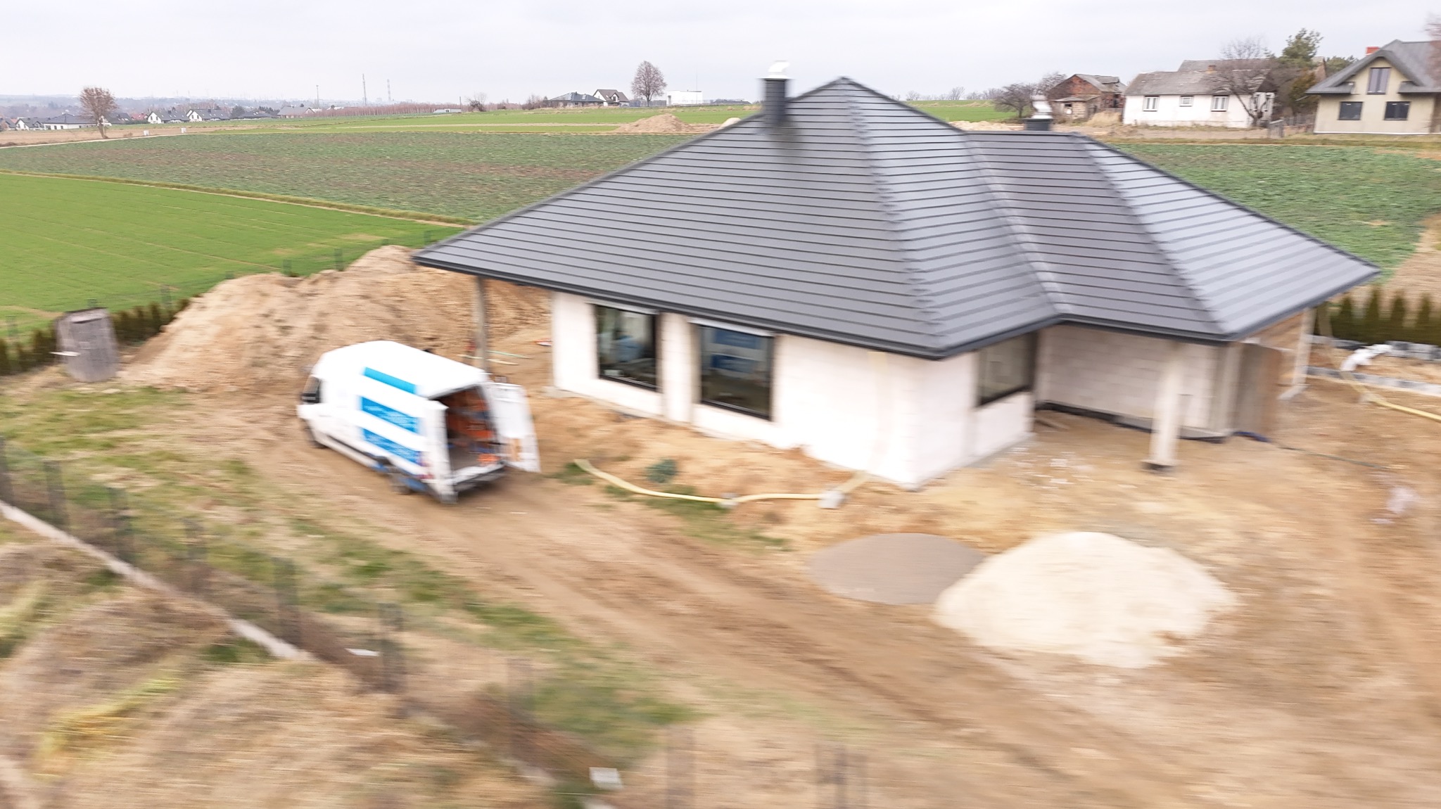 Niedokończony dom z szarą dachówką i białymi ścianami, z otwartym vanem serwisowym obok, na tle zielonych pól i zabudowań wiejskich.