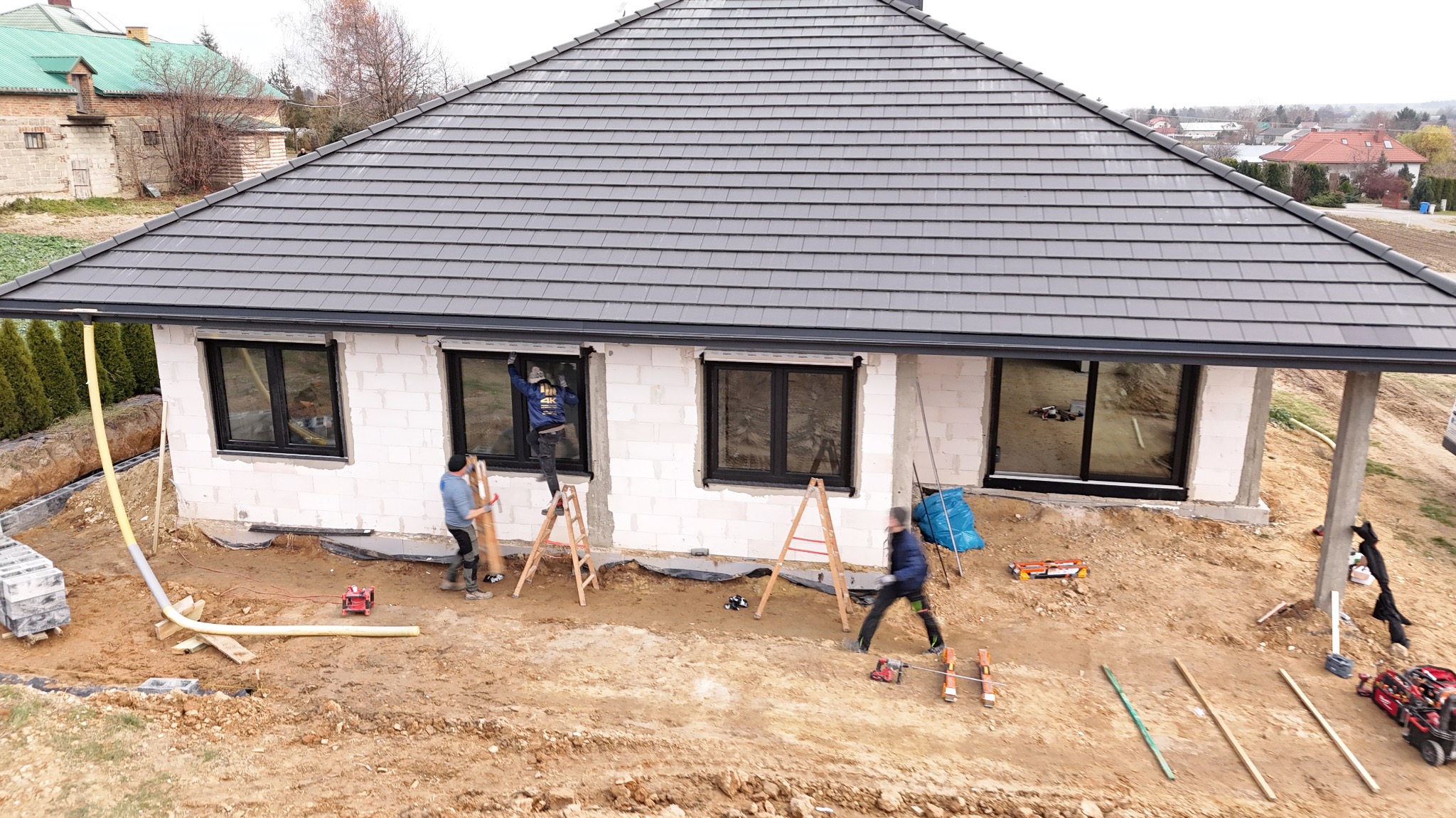Montaż okien z czarną ramą w surowym budynku. Trzech monterów przy pracy, drabiny, narzędzia. Widok z góry, na pierwszym planie ziemia.