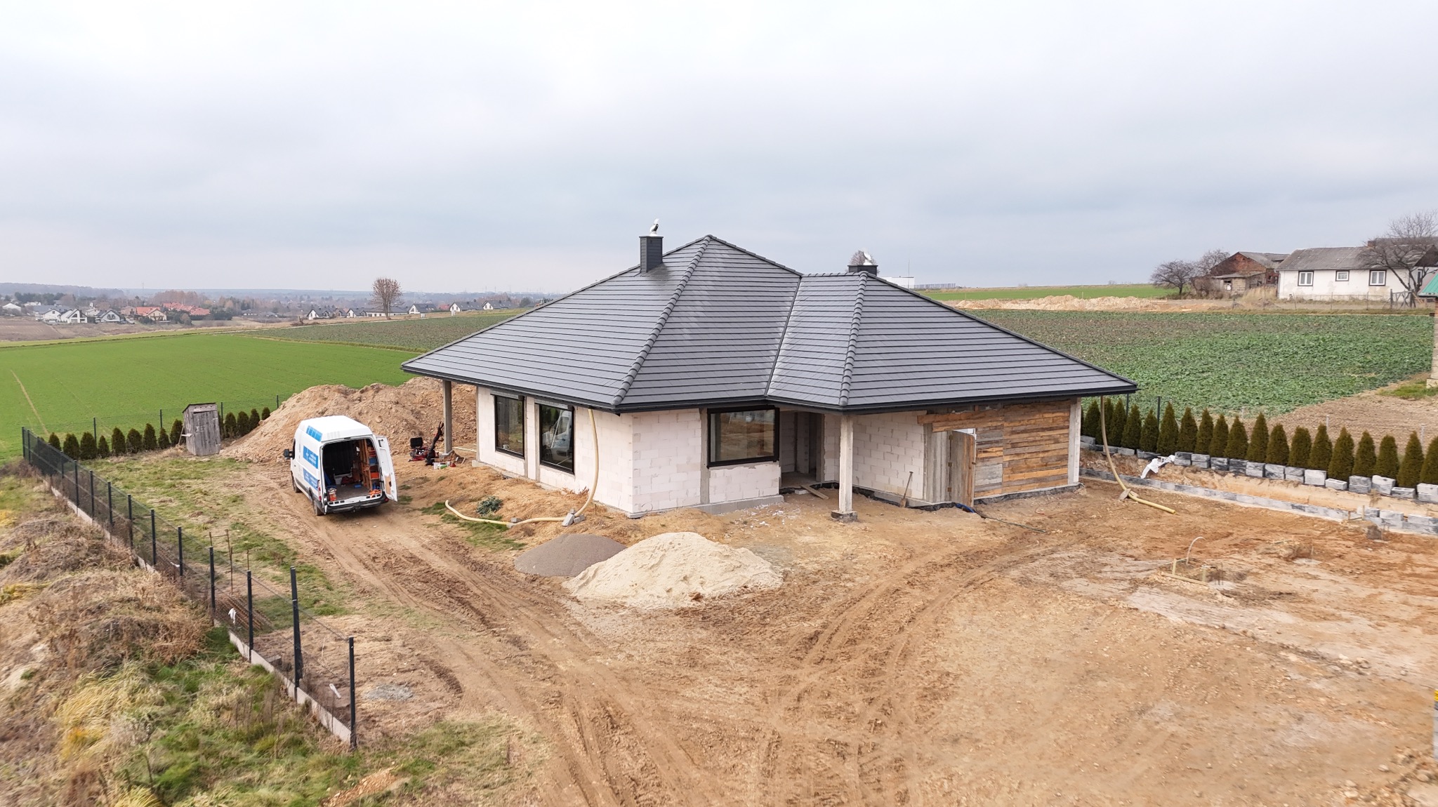 Dom jednorodzinny w trakcie budowy z czarną dachówką, widoczny z góry, z samochodem dostawczym na posesji i stertami piasku. Budowa w otoczeniu pól.