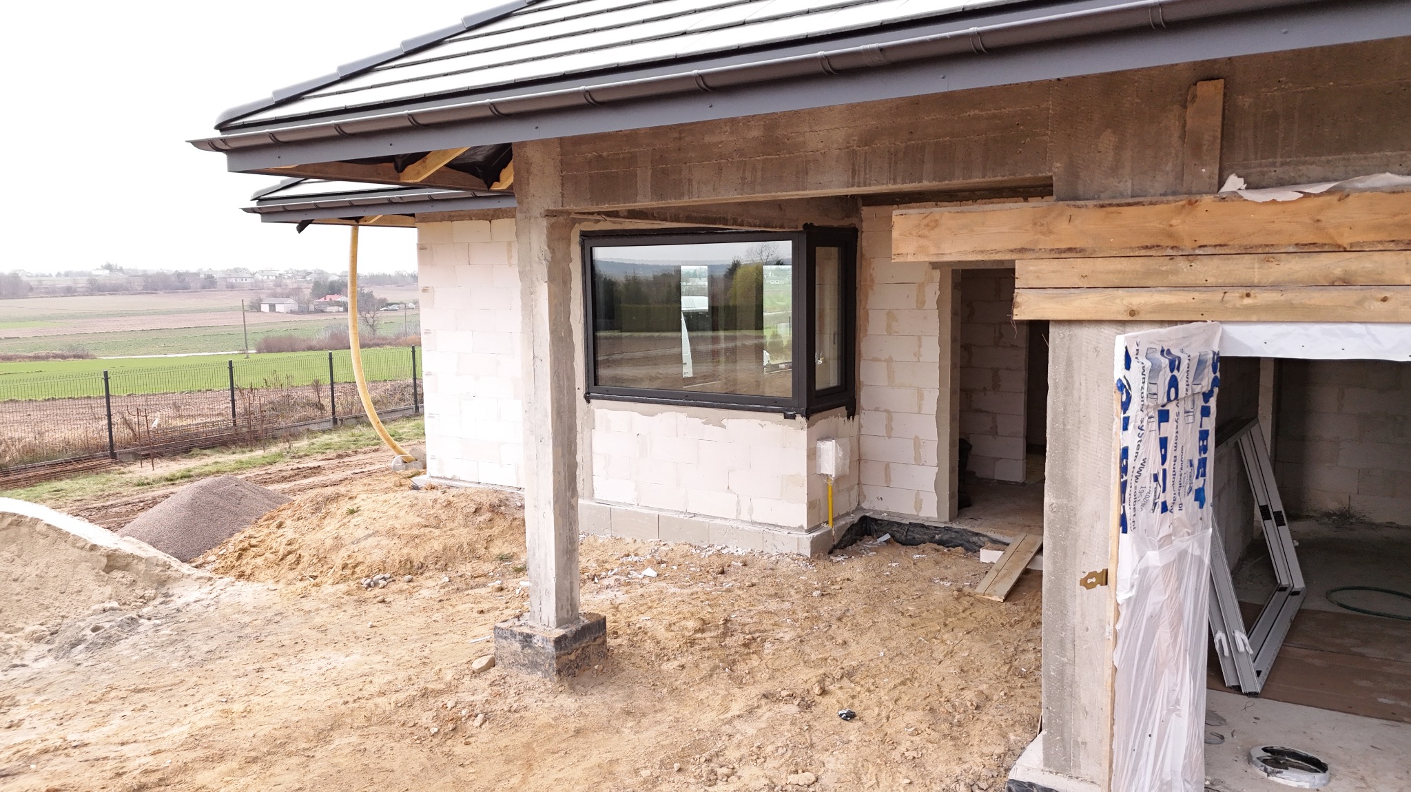 Częściowo zamontowane okno z czarną ramą w surowym domu z białych pustaków. Na zewnątrz widoczna ziemia i hałda piachu. Widok na pola.