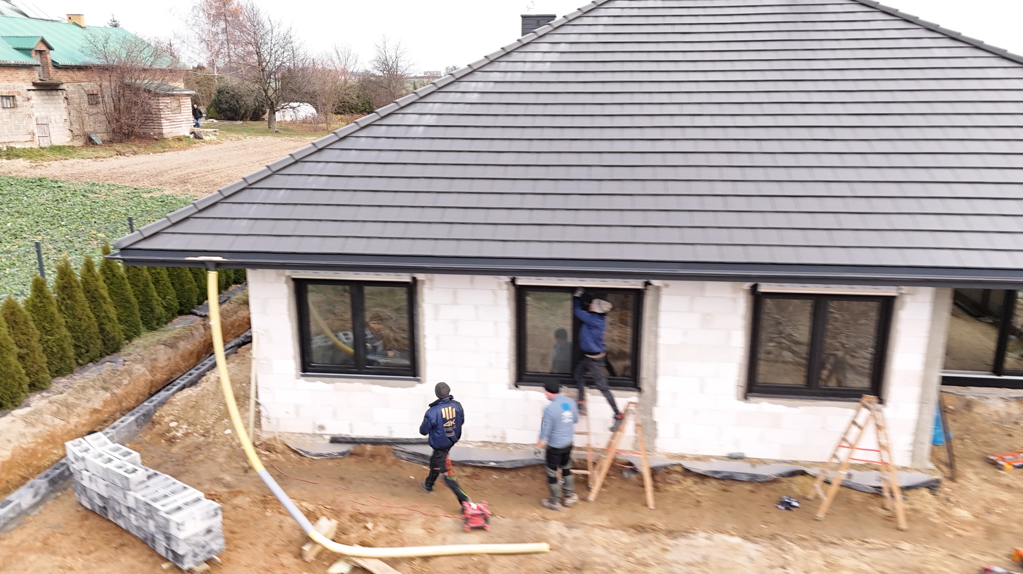 Montaż okien w surowym budynku z białego bloczka. Ekipa na drabinie instaluje okno. Widoczne czarne ramy okienne i szare dachówki.