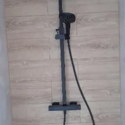 Czarny, matowy zestaw natryskowy z deszczownicą i słuchawką, zamontowany na ścianie wyłożonej płytkami imitującymi drewno.