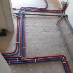 Instalacja wodna i grzewcza w trakcie montażu w surowym stanie, rury PEX czerwone i niebieskie połączone z szarymi rurami kanalizacyjnymi, mocowane do ściany za pomocą białych uchwytów.