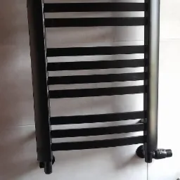 Czarny, drabinkowy grzejnik łazienkowy, zamontowany na jasnych, kwadratowych płytkach, z widocznym zaworem termostatycznym.