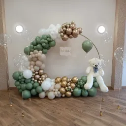 Dekoracja balonowa w kształcie półkola z pluszowym misiem, w kolorach zieleni, bieli i złota, na drewnianej podłodze, z napisem 'Oh Baby'.