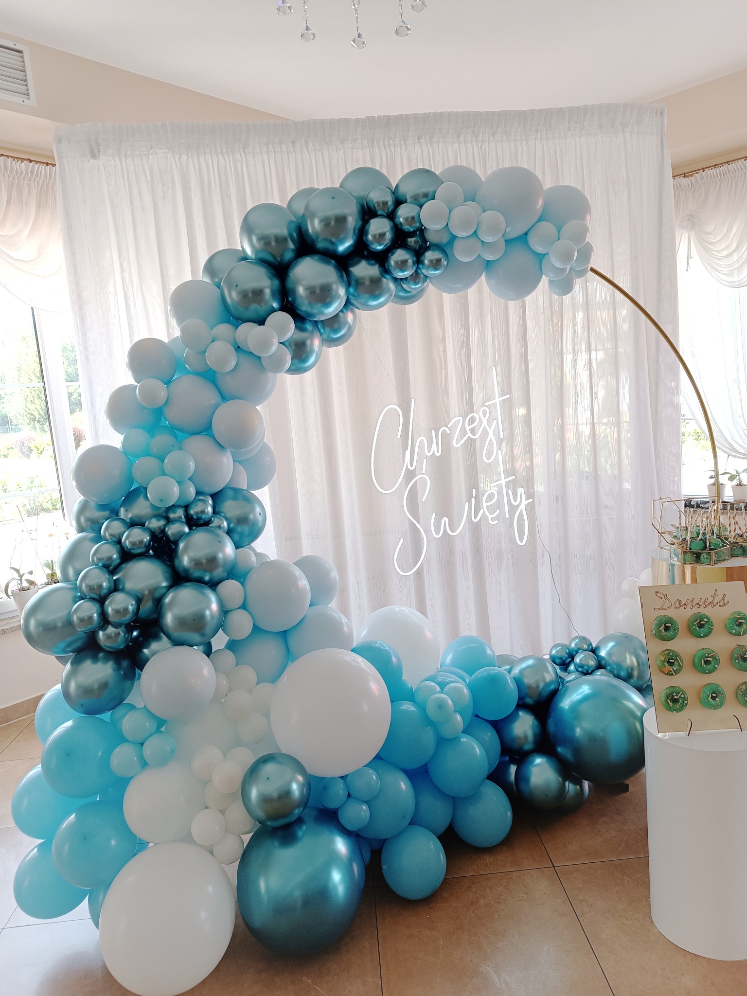 Dekoracja balonowa w odcieniach błękitu i bieli, tworząca łuk na przyjęcie, z napisem neonowym 'Chrzest Święty' i stojakiem z zielonymi pączkami.