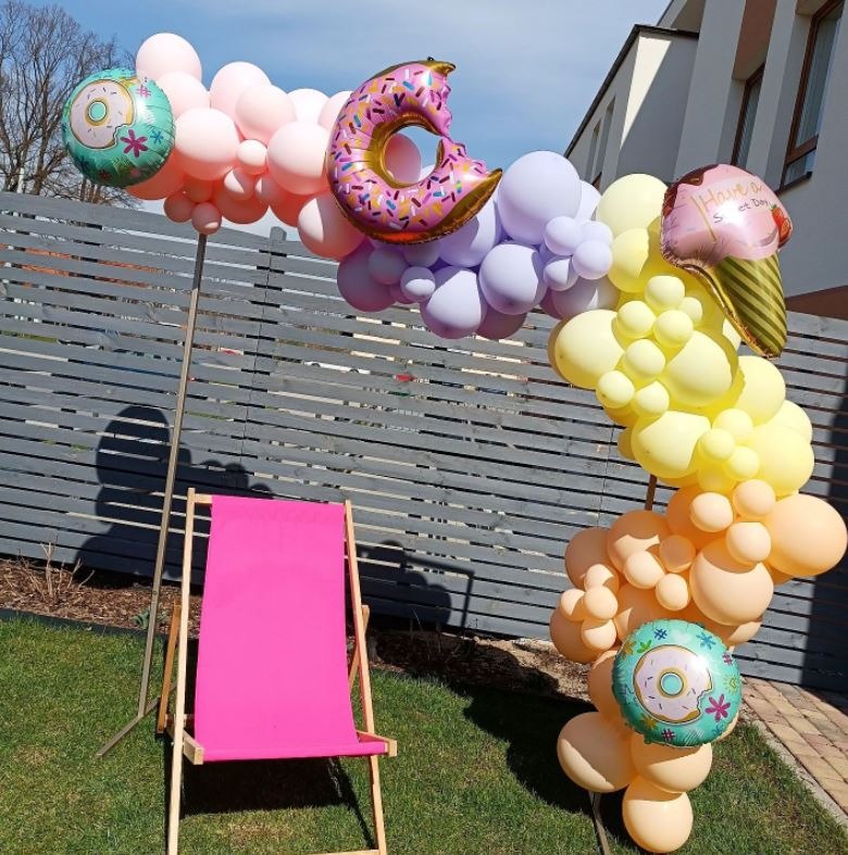 Dekoracja balonowa w kształcie łuku z motywami słodyczy, ustawiona na trawniku obok różowego leżaka.