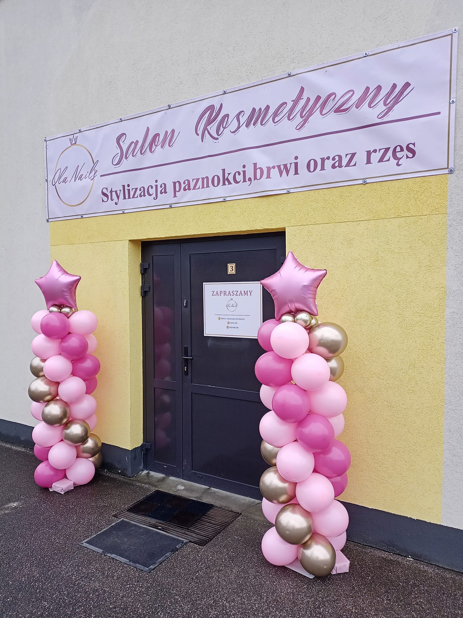 Wejście do salonu kosmetycznego udekorowane kolumnami z różowych i złotych balonów zwieńczonych gwiazdami, nad drzwiami szyld reklamowy z napisem 'Salon Kosmetyczny Stylizacja paznokci, brwi...