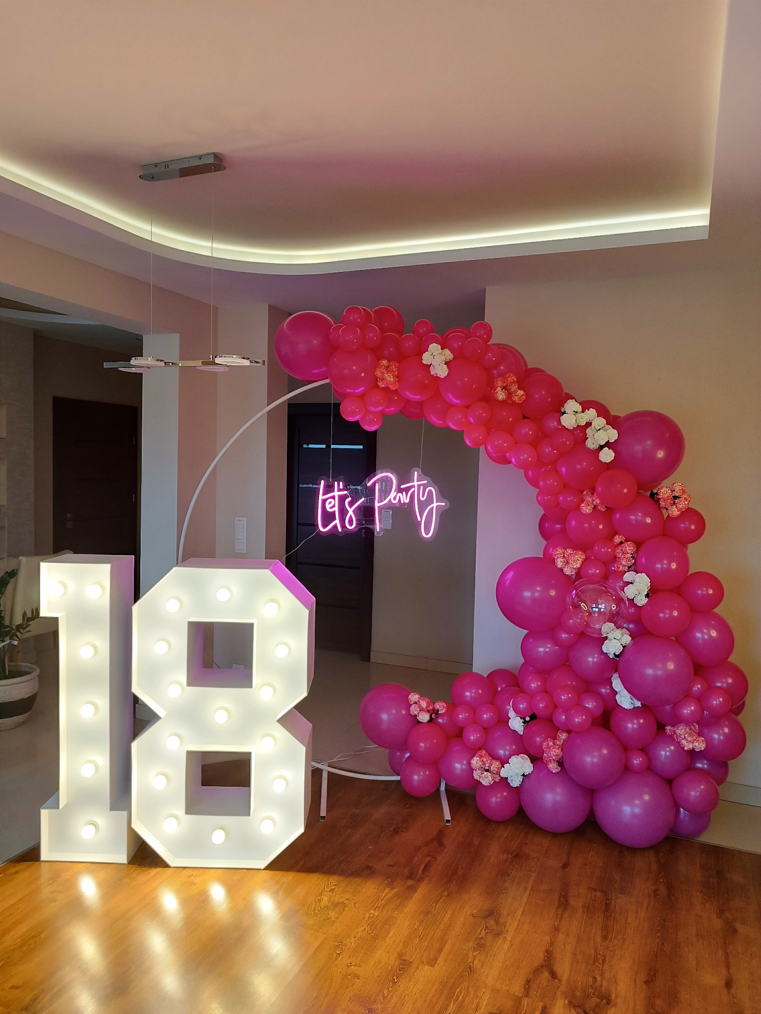 Dekoracja na 18 urodziny: podświetlane cyfry '18' i łuk z różowych balonów z białymi kwiatami, neon 'Let's Party' w tle, nowoczesne oświetlenie sufitowe.