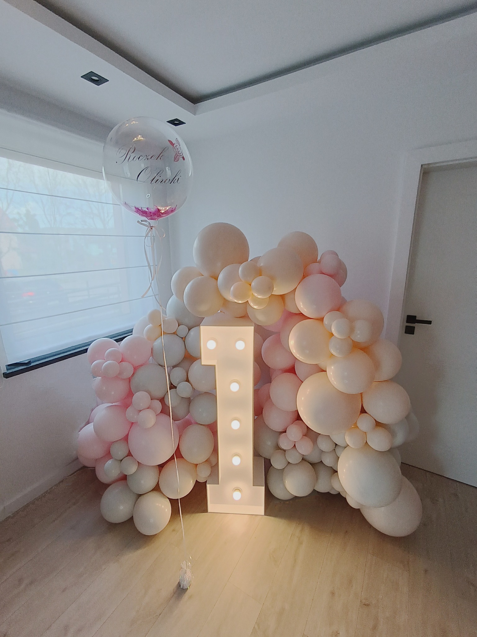 Dekoracja balonowa w odcieniach różu i bieli z podświetlaną cyfrą jeden, przeznaczona na roczek dziecka, z dodatkowym balonem z napisem 'Roczek Oliwi'