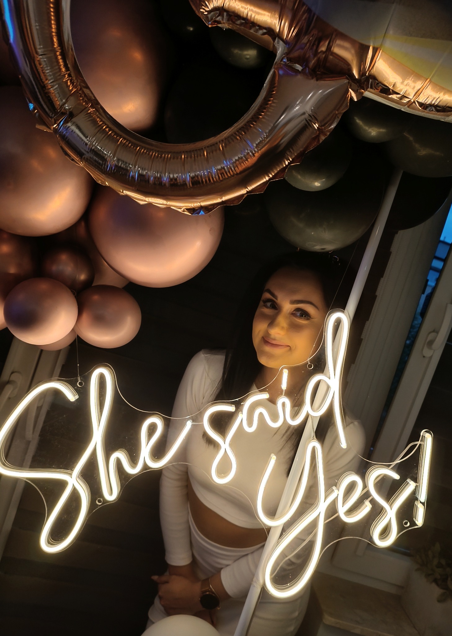 Uśmiechnięta kobieta w białym stroju pozuje z neonowym napisem 'She said yes!' na tle balonów w odcieniach różu i czerni.