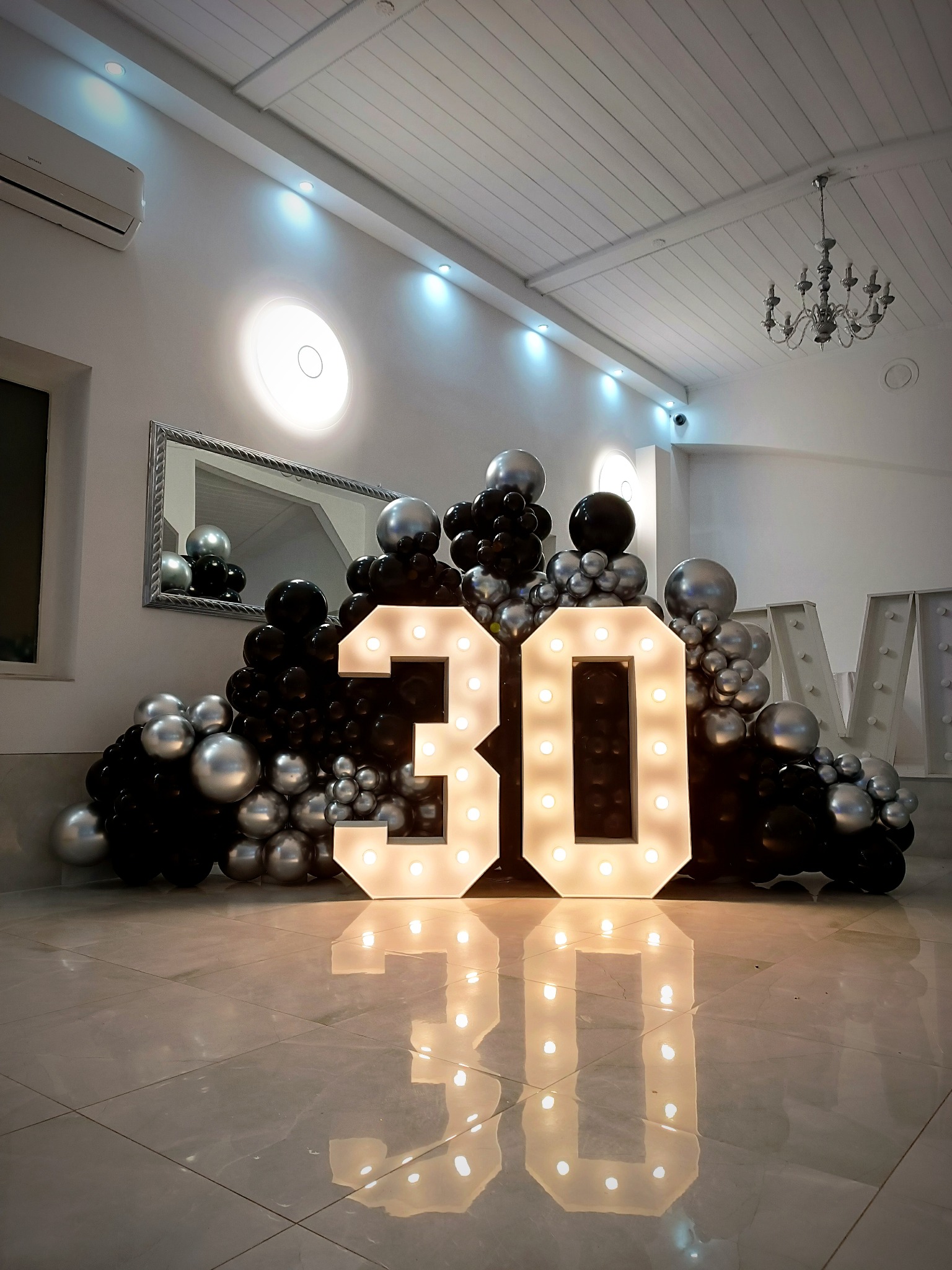 Oświetlona dekoracja urodzinowa z liczbą 30 otoczoną czarno-srebrnymi balonami na błyszczącej podłodze, widoczny fragment dekoracji literowej w tle.