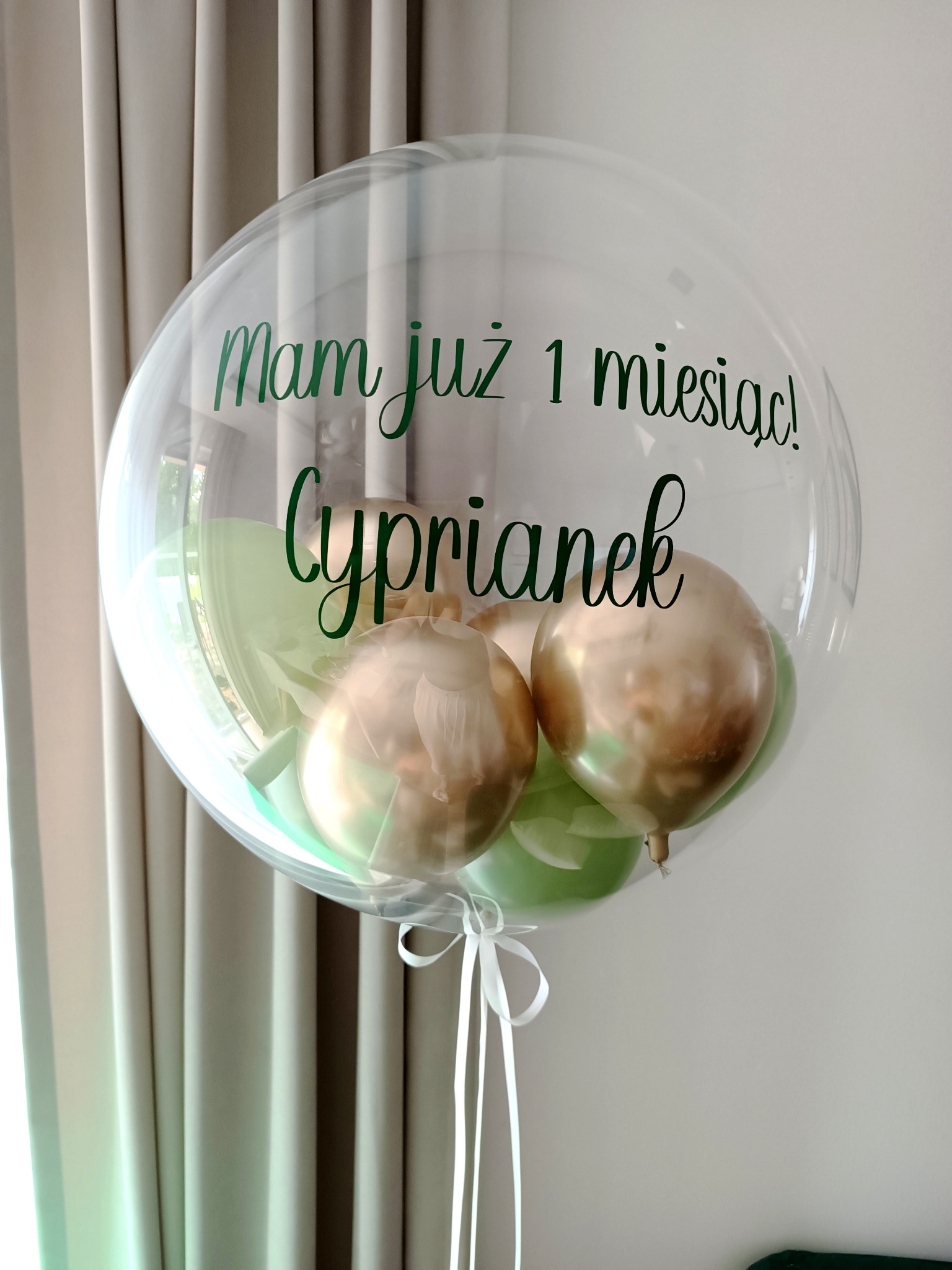 Przezroczysty balon z napisem 'Mam już 1 miesiąc! Cyprianek' w kolorze zielonym, wypełniony małymi balonami w kolorze złotym i pastelowej zieleni, zawiązany białą wstążką, tło stanowią beżowe zasłony.