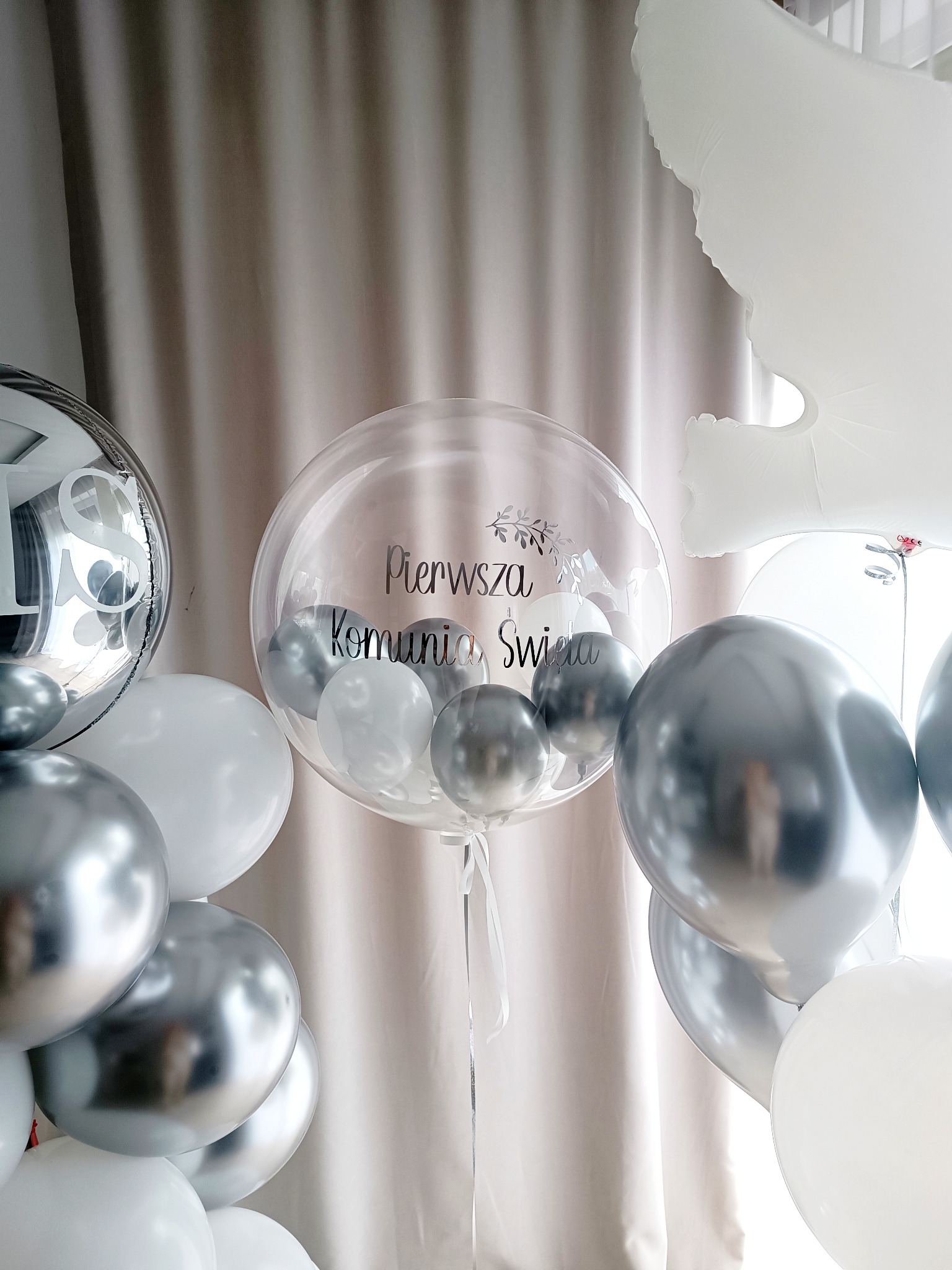 Dekoracja balonowa w odcieniach bieli i srebra z napisem 'Pierwsza Komunia Święta' na przezroczystym balonie.