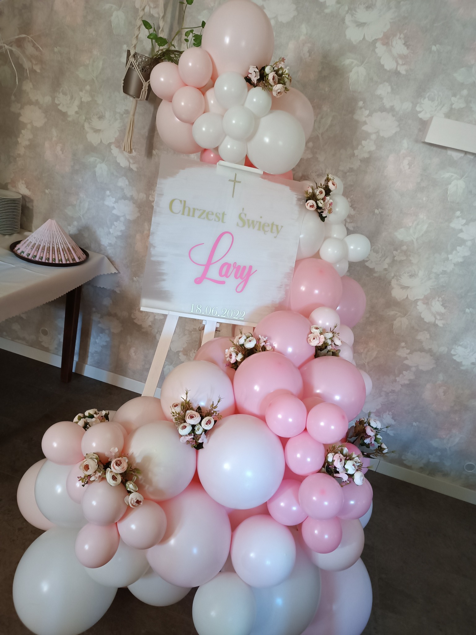 Dekoracja balonowa w odcieniach różu i bieli z drobnymi kwiatami, ustawiona obok tablicy na sztaludze z napisem 'Chrzest Święty Lary' i datą 18.06.2022, na tle ściany w kwiaty.