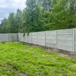 Długi, szary płot betonowy z poziomymi panelami, oddzielający trawnik od zieleni drzew. Widoczne słupki i łączenia paneli. Tło: las i fragment domu.