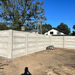 Rem-Bet - Nowoczesne ogrodzenie betonowe z poziomymi panelami, widoczne z zewnątrz posesji, z shadow osoby robiącej zdjęcie. Błękitne niebo i zieleń drzew w tle.