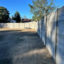 Szary, prefabrykowany płot betonowy w słoneczny dzień. Widoczne ślady opon na ziemi i fragmenty budynków w tle. Ujęcie z perspektywy.