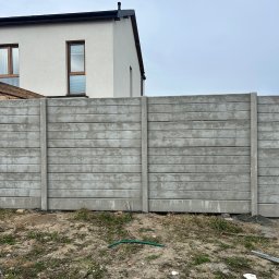 Rem-Bet - Szary, prefabrykowany płot betonowy z poziomymi panelami na nierównym terenie, z fragmentem białego domu w tle pod pochmurnym niebem. Widoczne elementy montażowe.