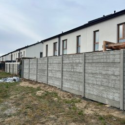 Rem-Bet - Szary, prefabrykowany płot betonowy z poziomymi żłobieniami, oddzielający szereg domów z jasną elewacją i brązowymi oknami, na tle pochmurnego nieba.