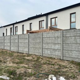Rem-Bet - Długi, szary mur z prefabrykatów betonowych oddziela szereg domów z jasną elewacją i brązowymi oknami. Widoczne elementy drewnianej konstrukcji za murem.