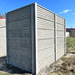 Rem-Bet Ogrodzenia - Fragment nowo postawionego, szarego ogrodzenia betonowego imitującego cegłę, na tle błękitnego nieba i zielonej trawy. Widoczne ślady prac ziemnych.