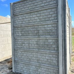 Rem-Bet Ogrodzenia - Fragment betonowego ogrodzenia z fakturą cegły, widoczne panele i słupki, surowy beton, błękitne niebo w tle. Ujęcie z bliska, podkreślające detale.