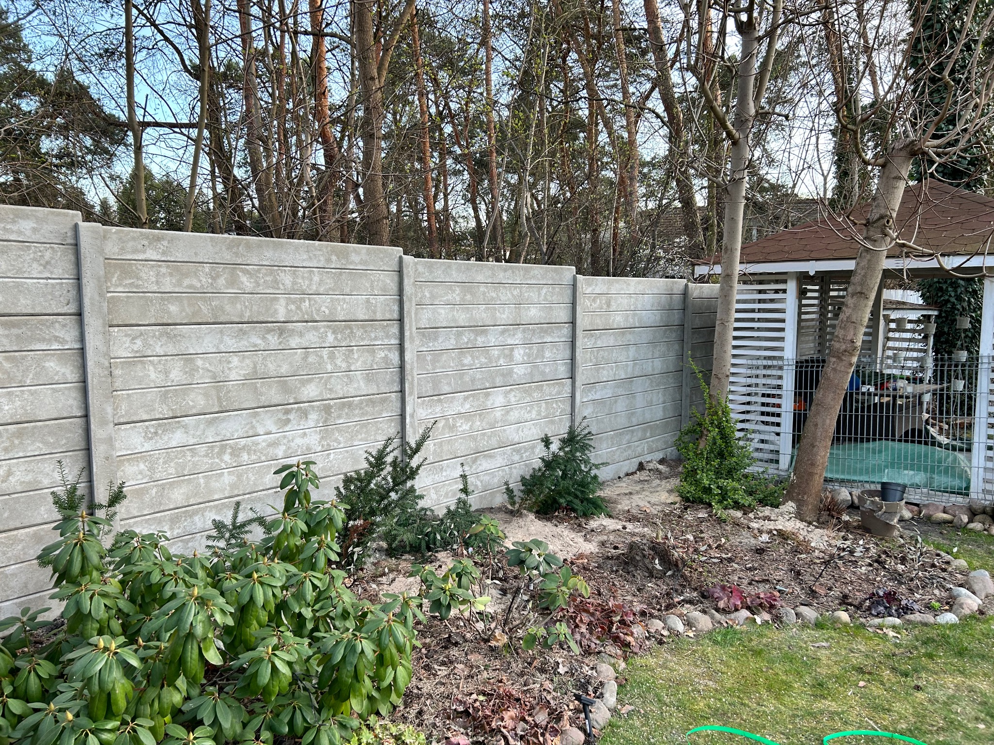 Szary, prefabrykowany płot z betonu architektonicznego, dzielący ogród z młodymi roślinami iglastymi i liściastymi, w tle altana i drzewa bezlistne.