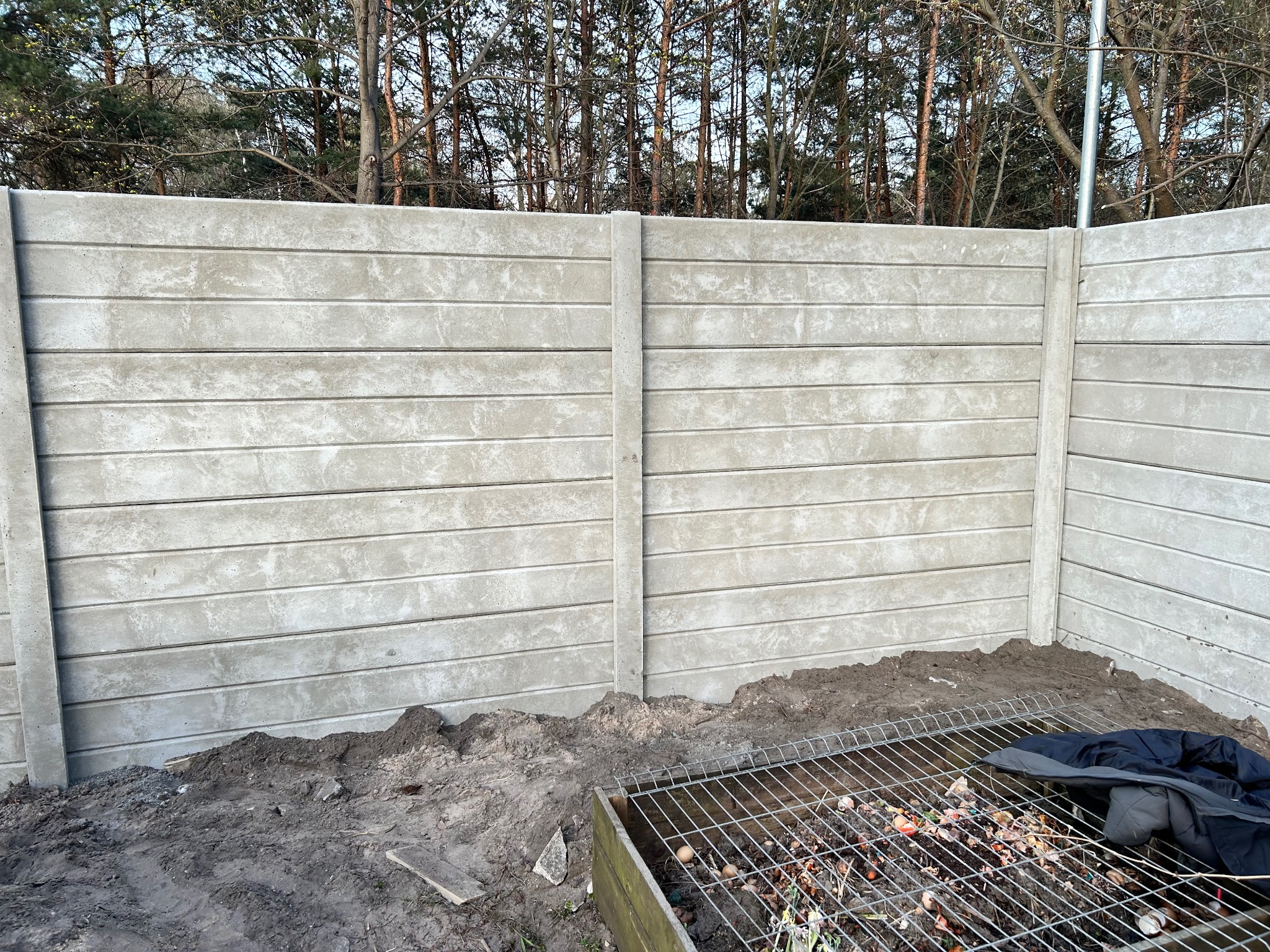 Nowo postawiony, szary, betonowy płot panelowy z widocznym fragmentem kompostownika z resztkami organicznymi i kurtką na wierzchu, na tle drzew.