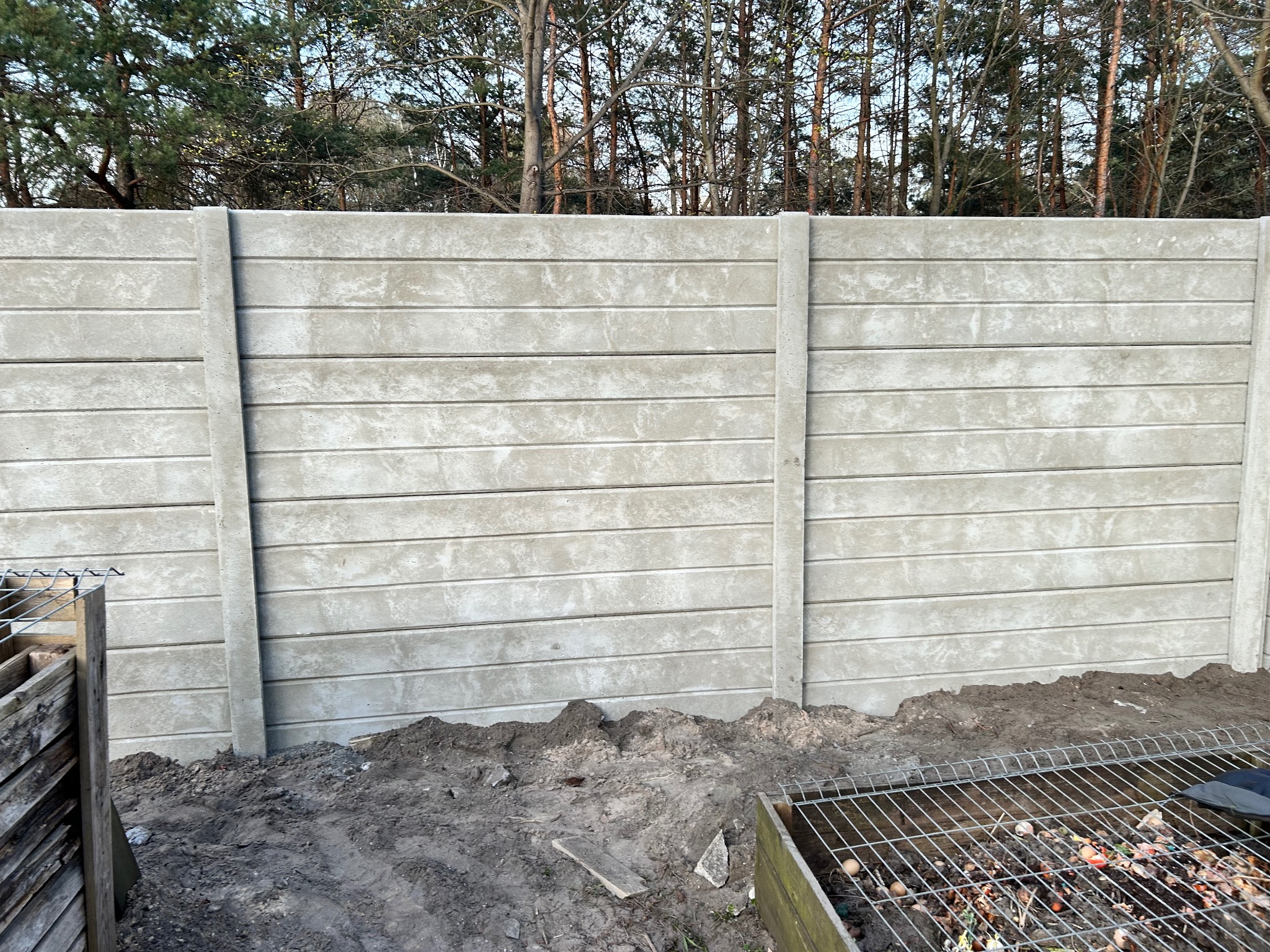 Szary, prefabrykowany płot z betonu, z widocznymi poziomymi panelami i pionowymi słupkami, w tle drzewa bez liści, ziemia wokół płotu naruszona, widoczny kompostownik.