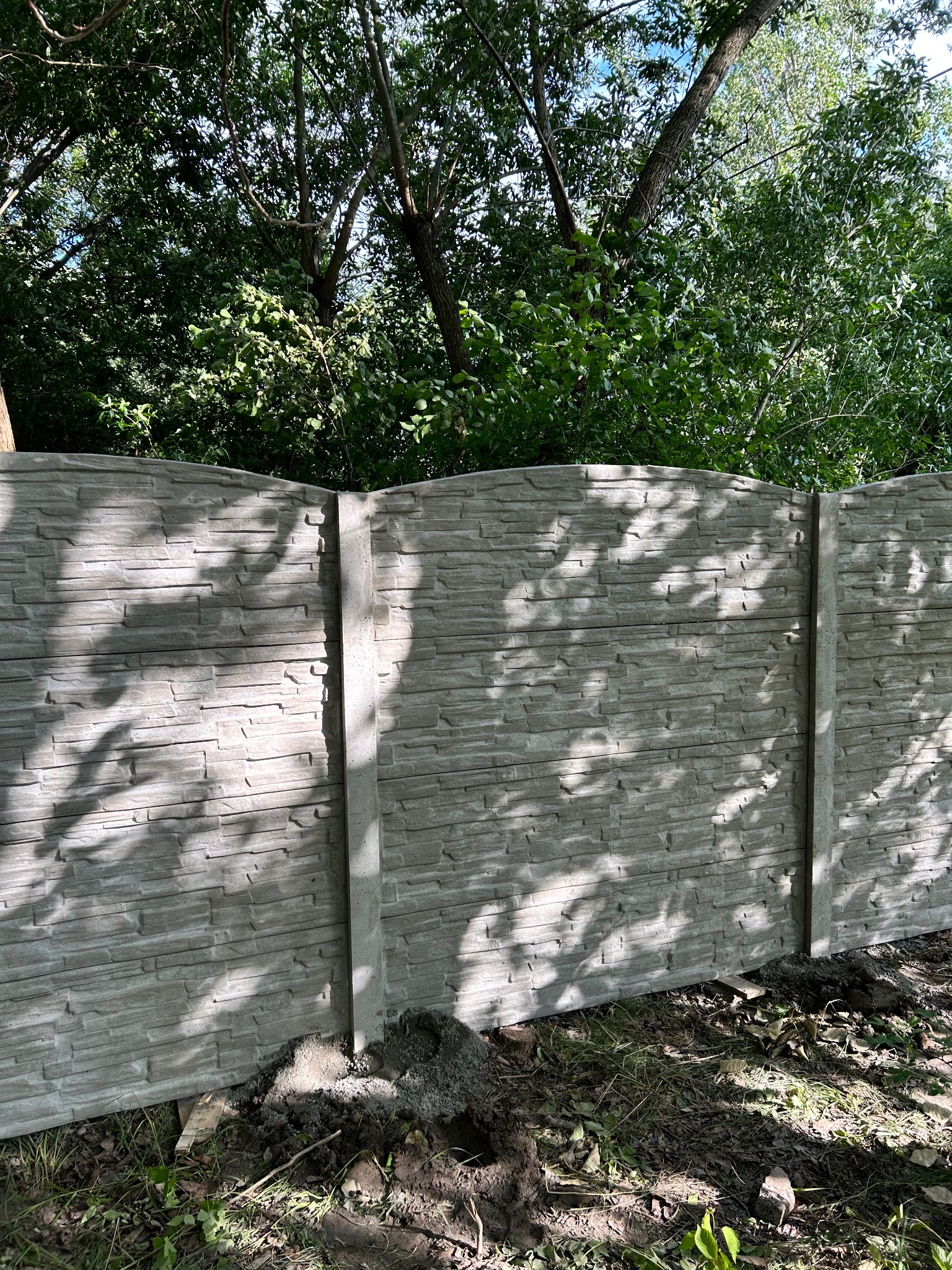 Fragment ogrodzenia betonowego z fakturą kamienia, oświetlonego przez słońce, z widocznym tłem zieleni drzew. Ujęcie z bliska, podkreślające detale.