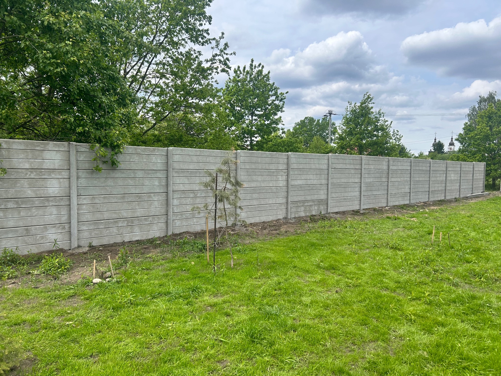 Długi, szary, prefabrykowany płot betonowy z poziomymi panelami, widoczny na tle zielonego trawnika i drzew. W tle majaczą wieże kościoła.