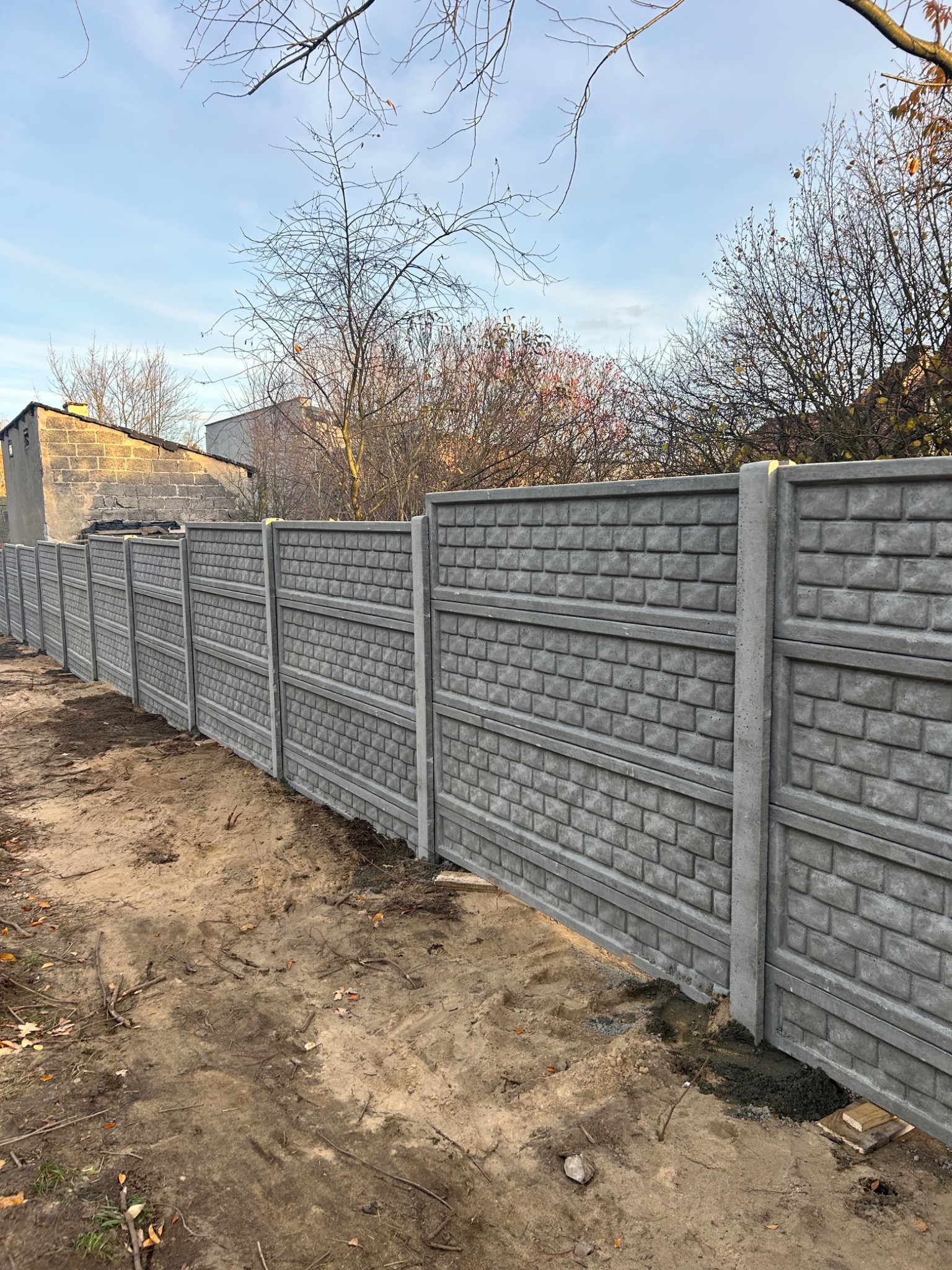 Długi, szary, betonowy płot z imitacją cegły, oddzielający posesje. Widoczne słupki i panele. Tło stanowią budynki i drzewa bez liści pod błękitnym niebem.