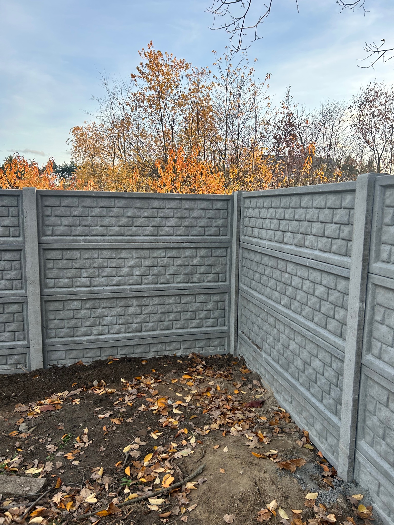 Fragment nowo postawionego, szarego ogrodzenia z prefabrykatów betonowych o fakturze cegły, z jesiennymi liśćmi u podstawy i drzewami w tle.