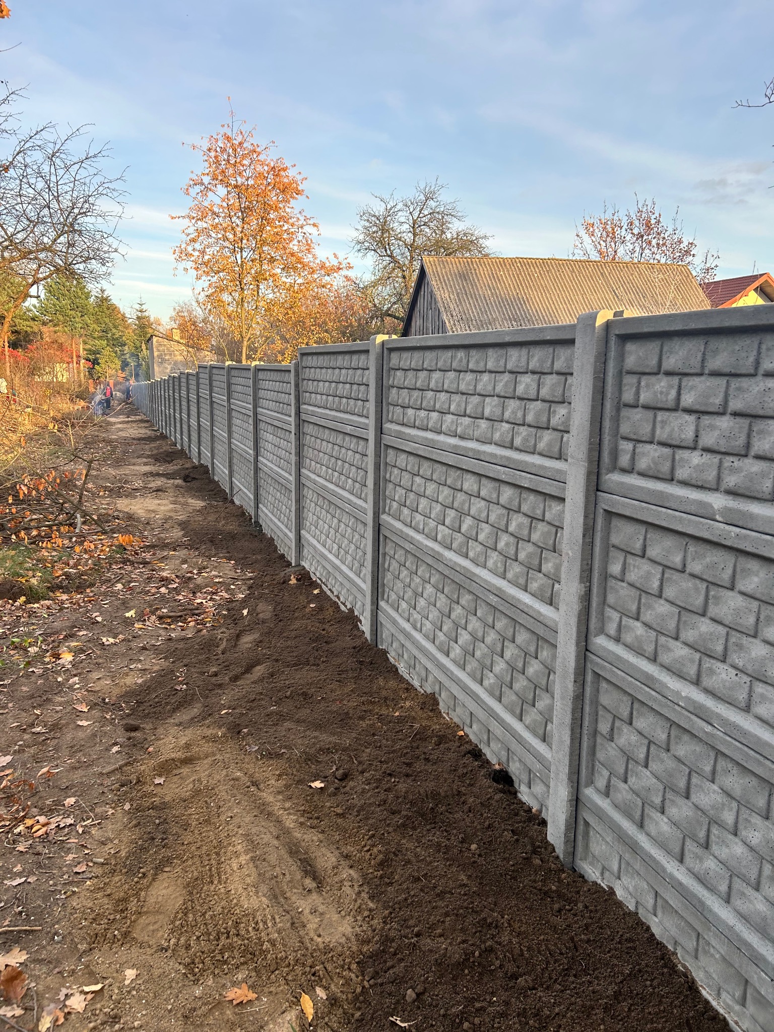 Długi, szary mur z prefabrykatów betonowych o fakturze cegły, ciągnący się wzdłuż działki. W tle drzewa i budynki, a w oddali pracownicy. Świeżo obsypana ziemia u podstawy.