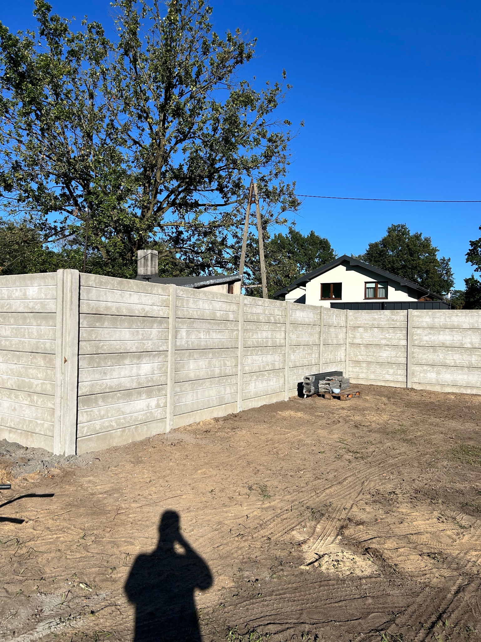 Nowoczesne ogrodzenie betonowe z poziomymi panelami, widoczne z zewnątrz posesji, z shadow osoby robiącej zdjęcie. Błękitne niebo i zieleń drzew w tle.