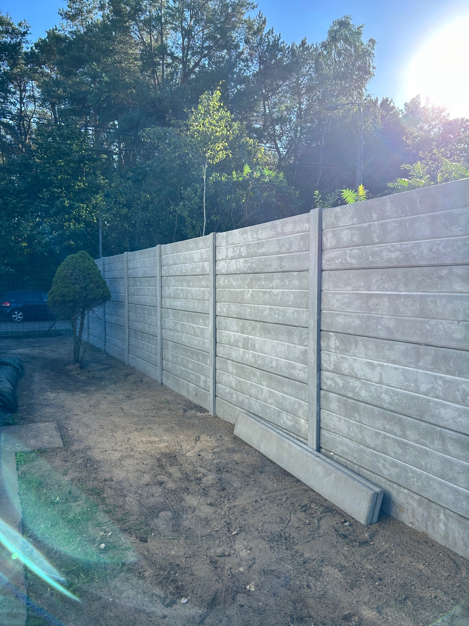 Szary, prefabrykowany płot betonowy z poziomymi panelami, częściowo zamontowany, z drzewem obok i widocznym samochodem w tle, w słoneczny dzień.