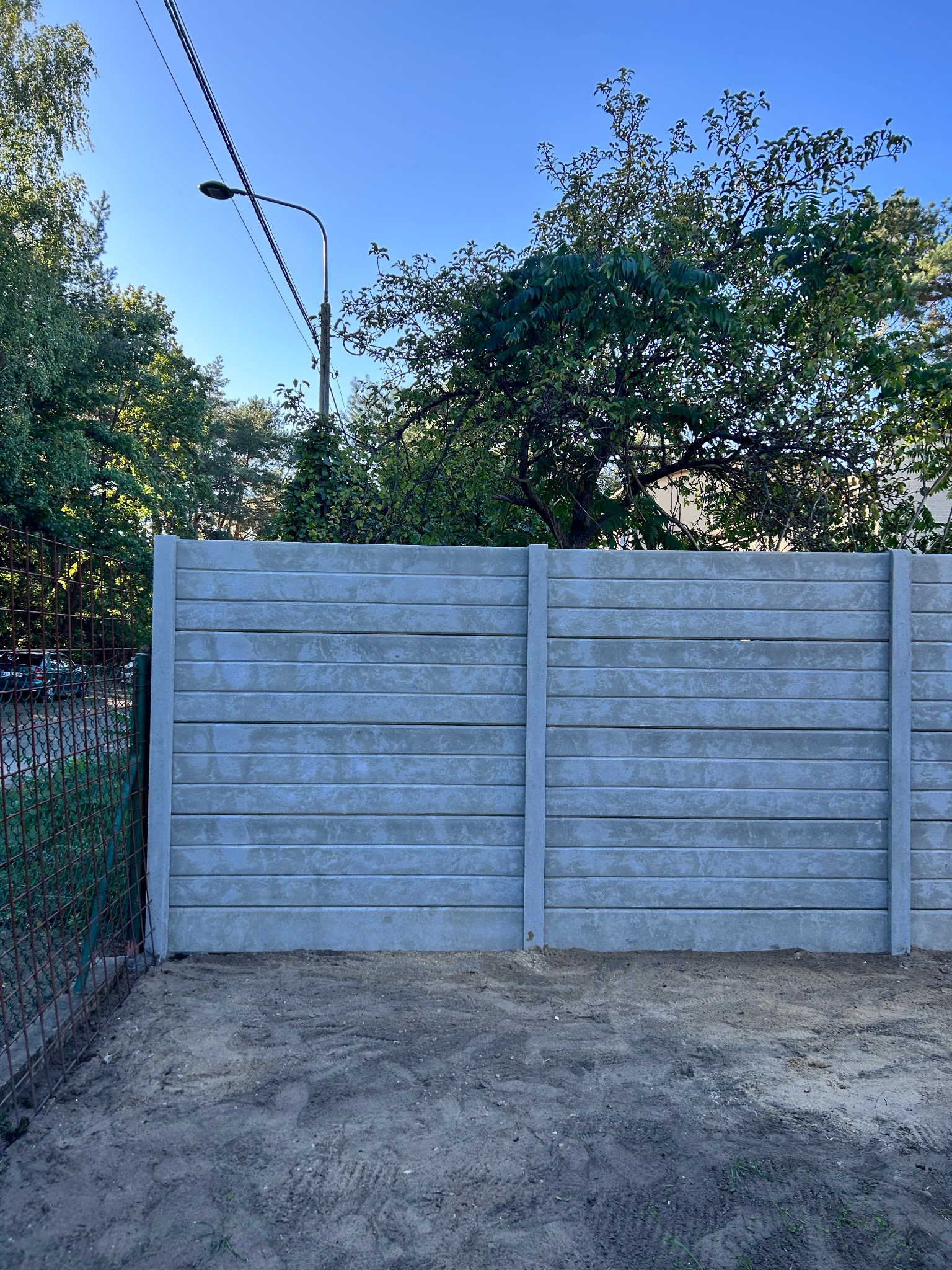 Szary, prefabrykowany płot betonowy z poziomymi panelami, widoczny z bliska, z zielenią w tle i fragmentem metalowej siatki po lewej stronie.