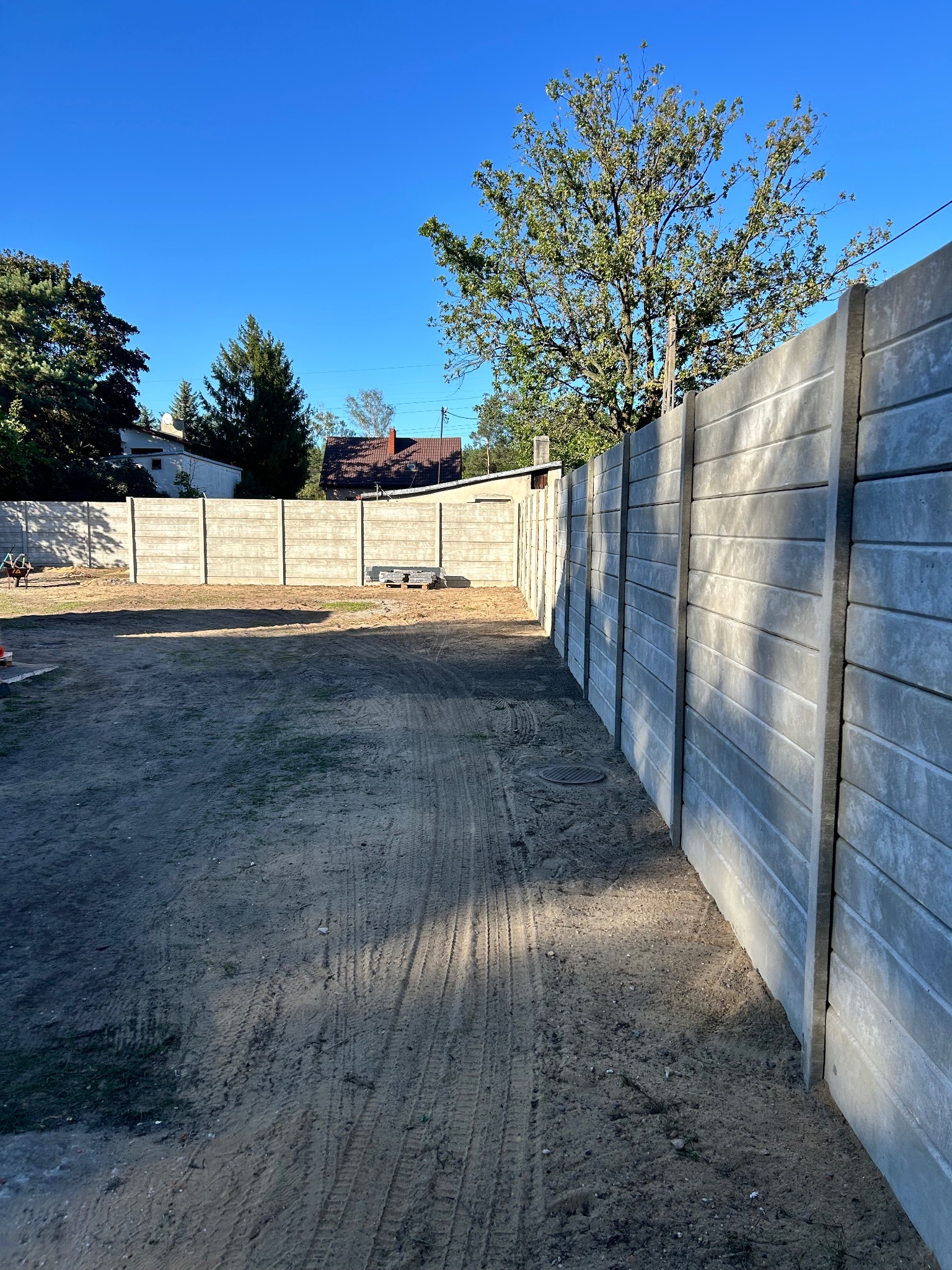 Szary, prefabrykowany płot betonowy w słoneczny dzień. Widoczne ślady opon na ziemi i fragmenty budynków w tle. Ujęcie z perspektywy.