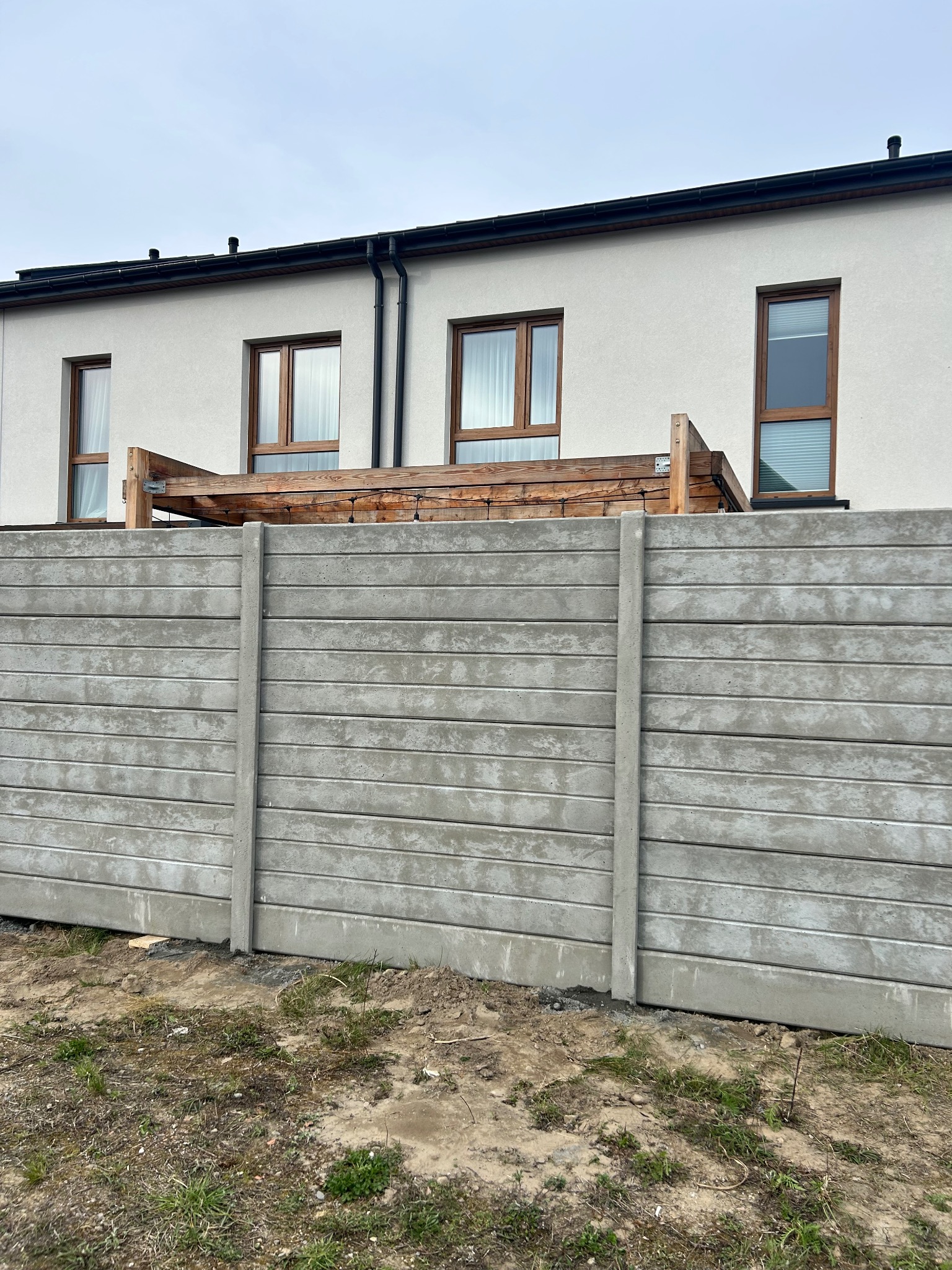 Szary, prefabrykowany płot betonowy z poziomymi panelami, widok z bliska na tle jasnego budynku z drewnianą konstrukcją na dachu. Surowy teren u podstawy.