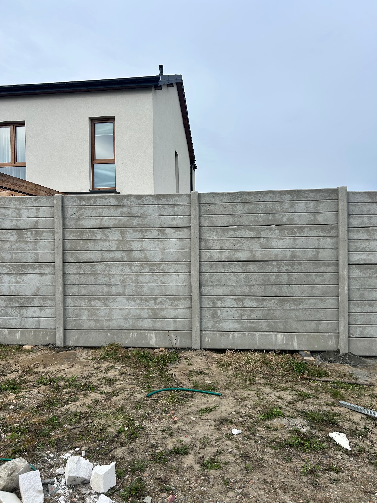 Szary, prefabrykowany płot betonowy z poziomymi panelami na nierównym terenie, z fragmentem białego domu w tle pod pochmurnym niebem. Widoczne elementy montażowe.