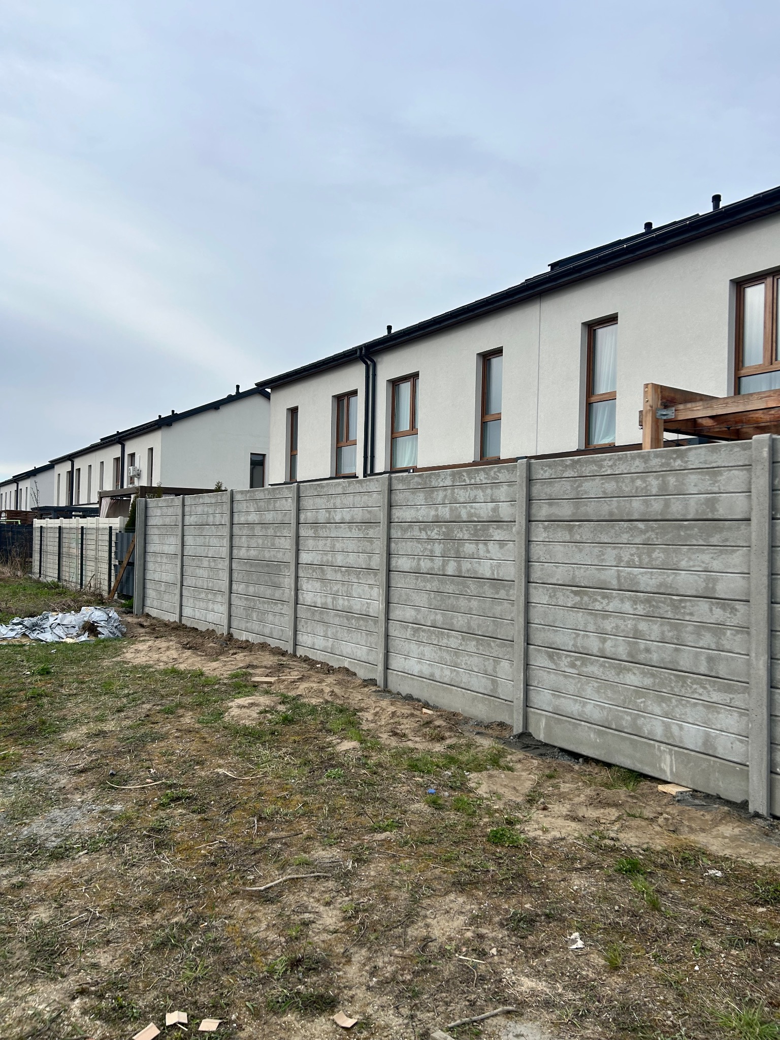 Szary, prefabrykowany płot betonowy z poziomymi żłobieniami, oddzielający szereg domów z jasną elewacją i brązowymi oknami, na tle pochmurnego nieba.