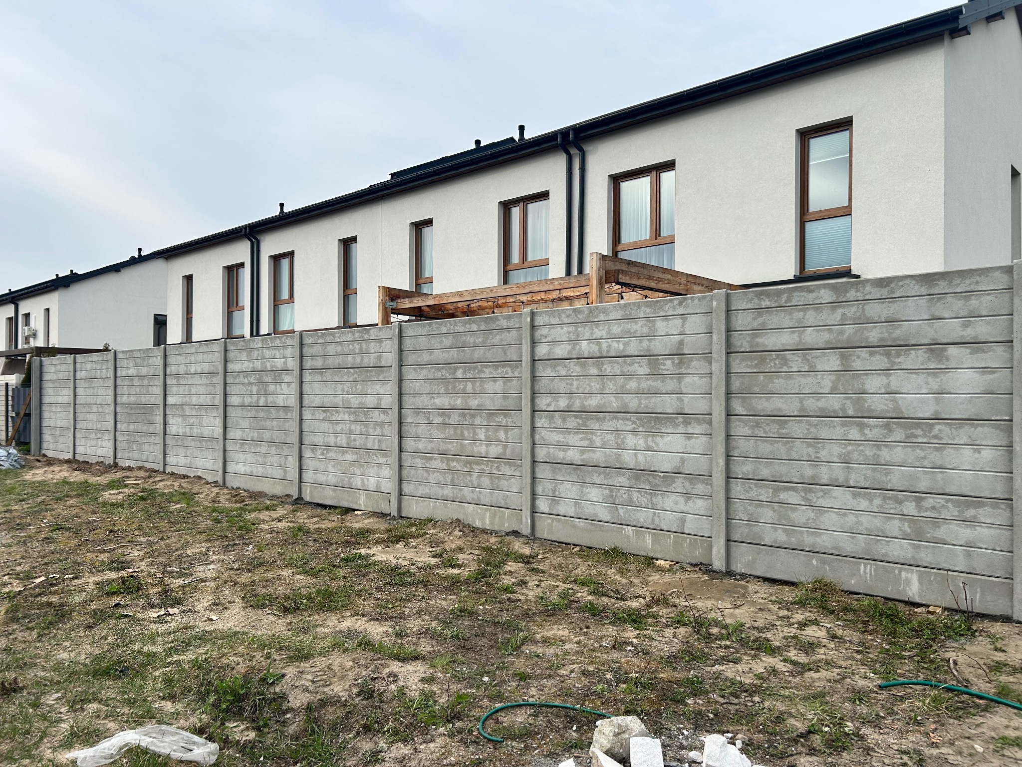 Długi, szary mur z prefabrykatów betonowych oddziela szereg domów z jasną elewacją i brązowymi oknami. Widoczne elementy drewnianej konstrukcji za murem.
