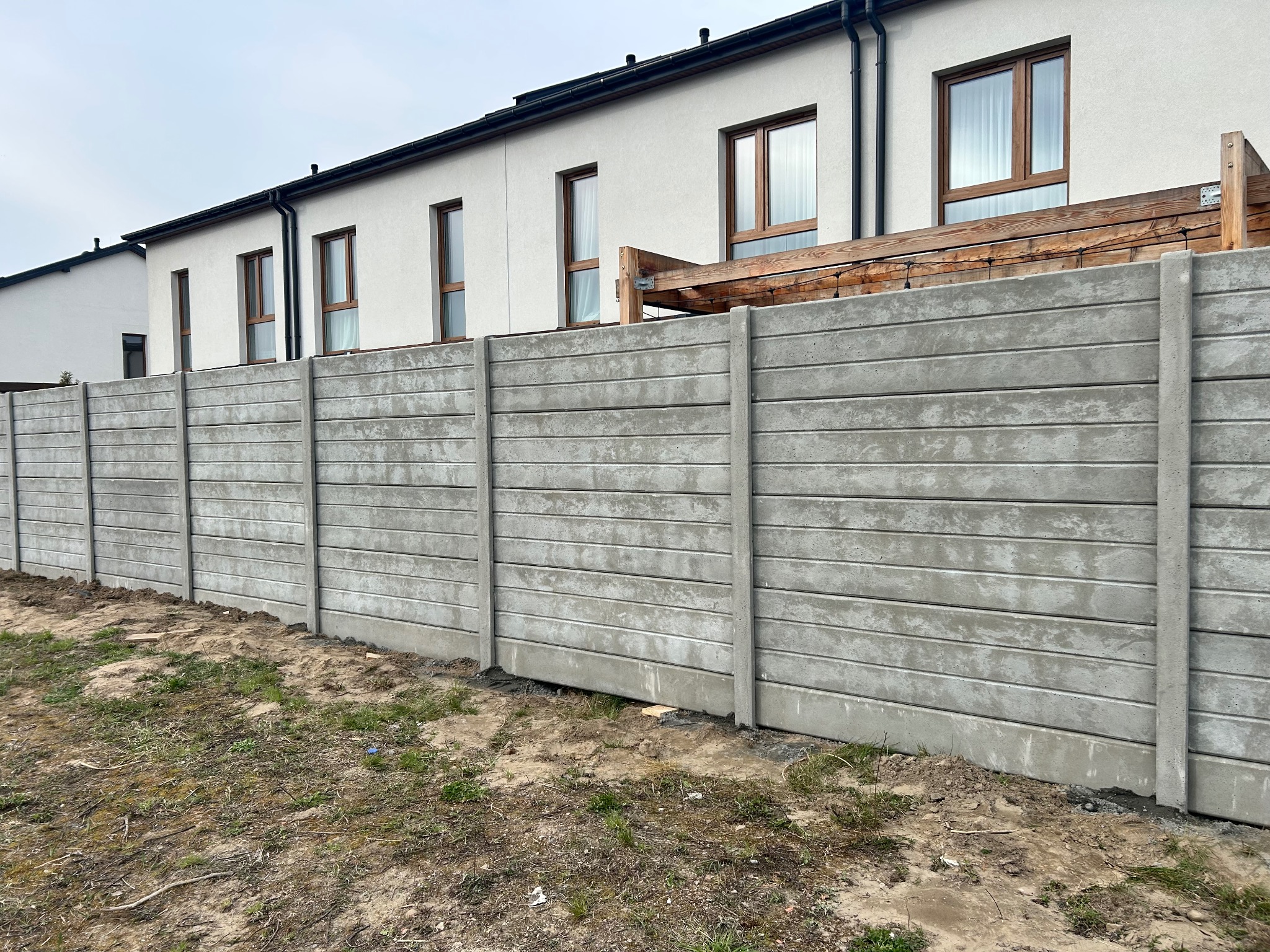 Szary, prefabrykowany płot betonowy z poziomymi panelami, oddzielający posesję z białym domem. Widoczne słupki i podmurówka. Trawa i ziemia u podstawy.