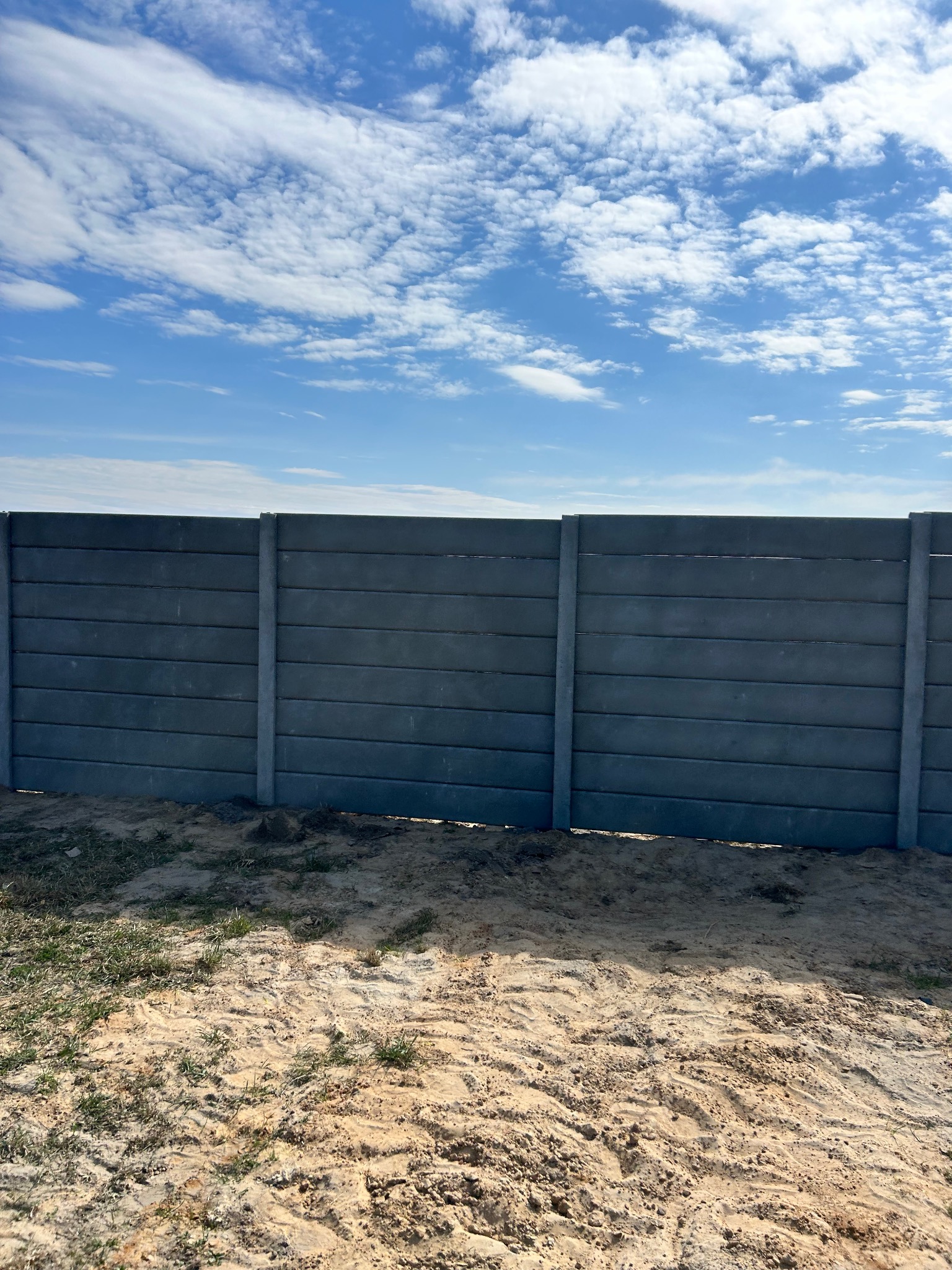Szary, prefabrykowany płot betonowy z poziomymi panelami na piaszczystym podłożu pod błękitnym niebem z chmurami. Widok z poziomu gruntu.