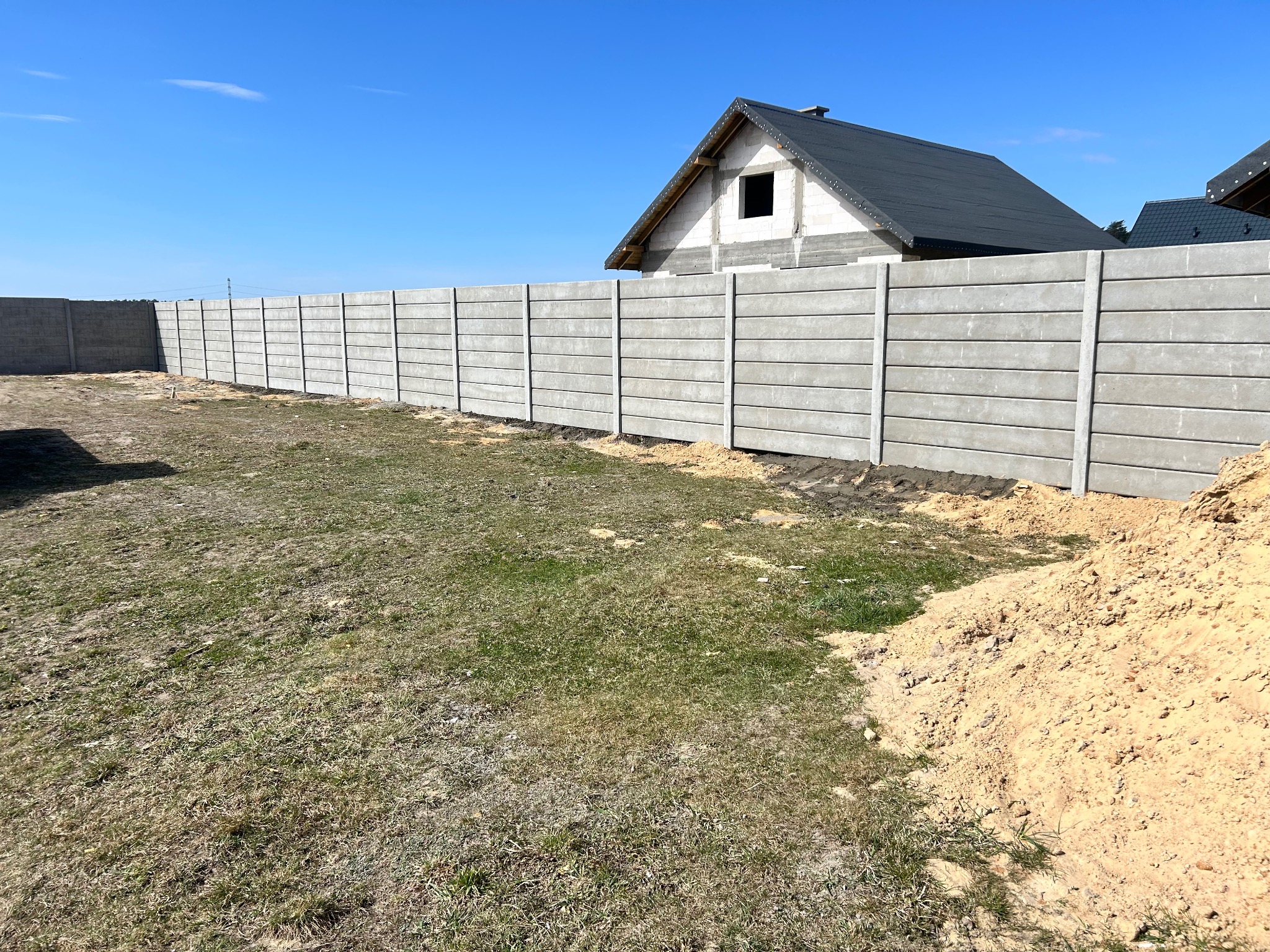 Długi, szary, prefabrykowany płot betonowy z poziomymi panelami, oddzielający posesję z budynkiem w stanie surowym, w słoneczny dzień.