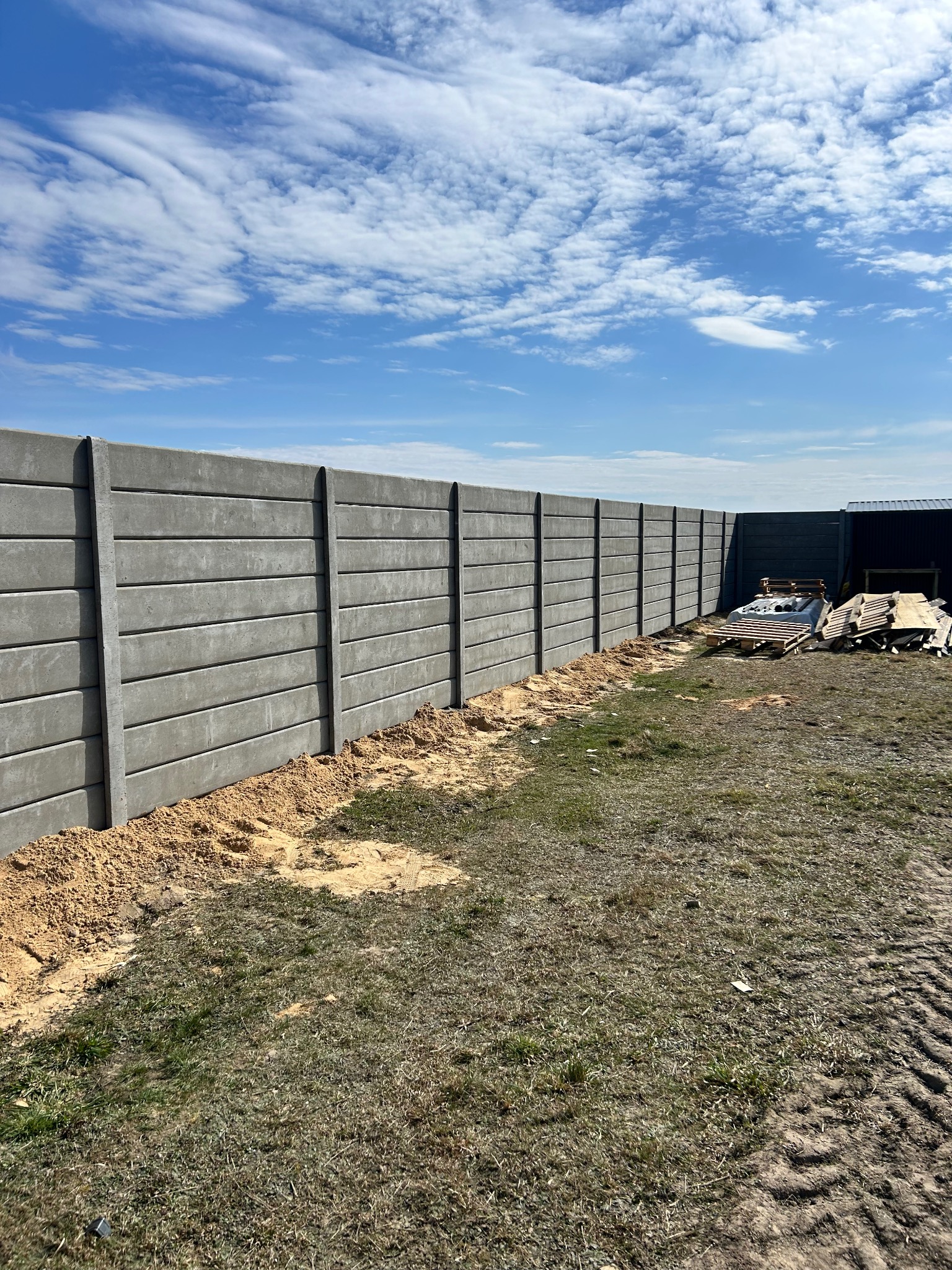 Długi, szary, betonowy płot panelowy z widocznymi poziomymi liniami łączeń, świeżo obsypany piaskiem, na tle błękitnego nieba z chmurami i zielonej trawy.