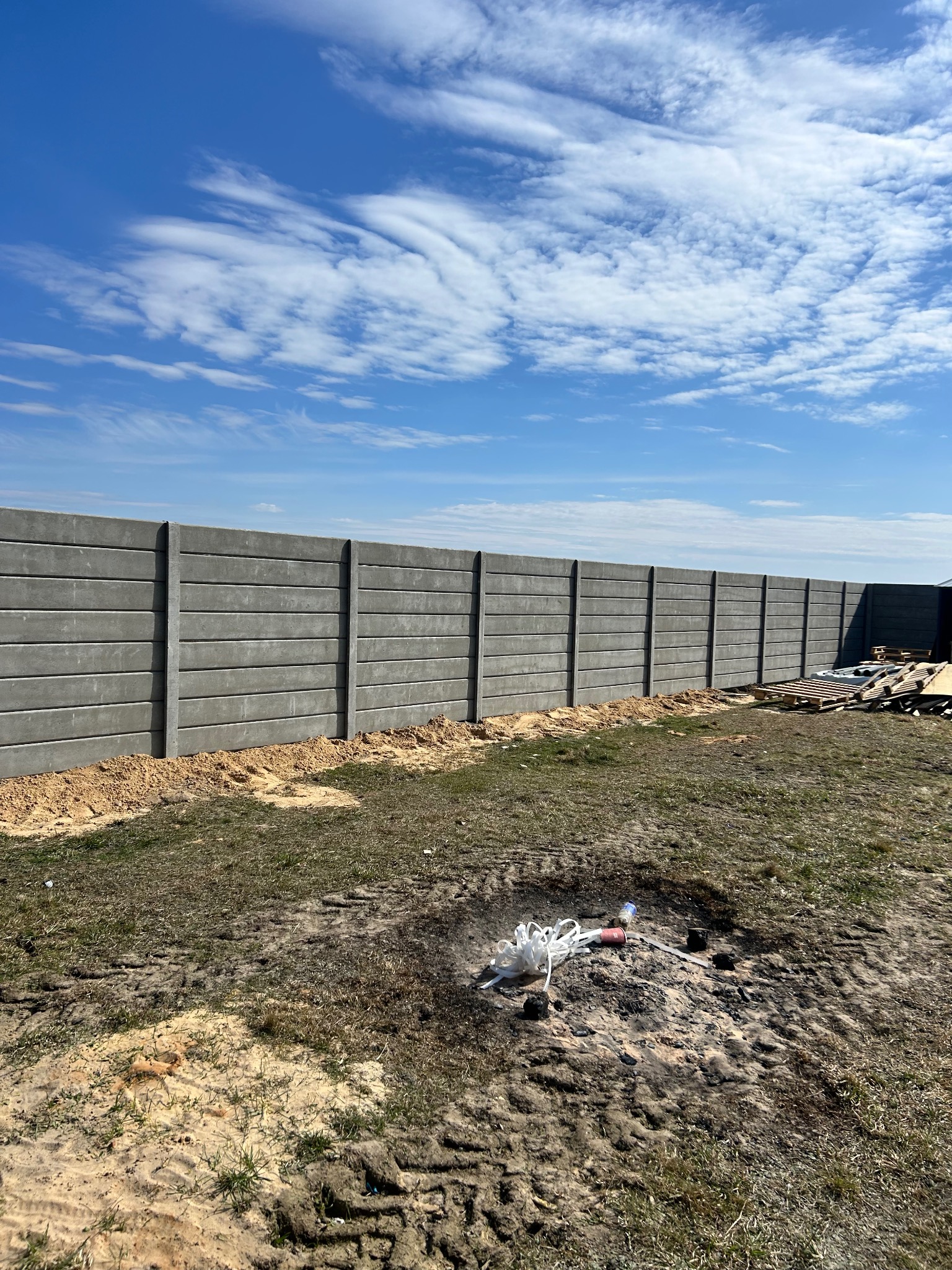 Długi, szary płot z prefabrykatów betonowych, widoczny w słoneczny dzień na tle błękitnego nieba z chmurami. Na pierwszym planie ziemia z resztkami po ognisku.